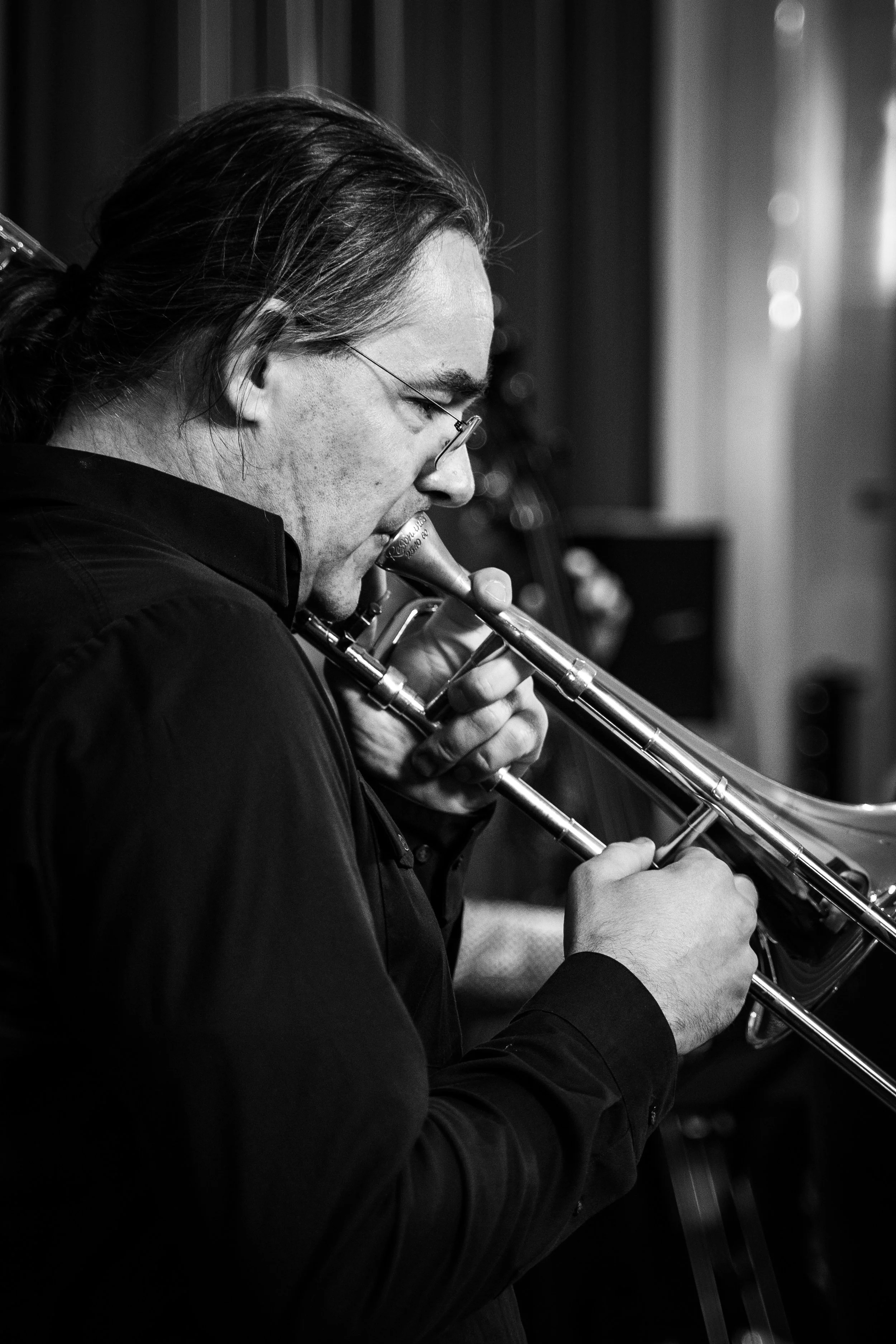 A black and white photograph of a man with long hair playing a trumpet.