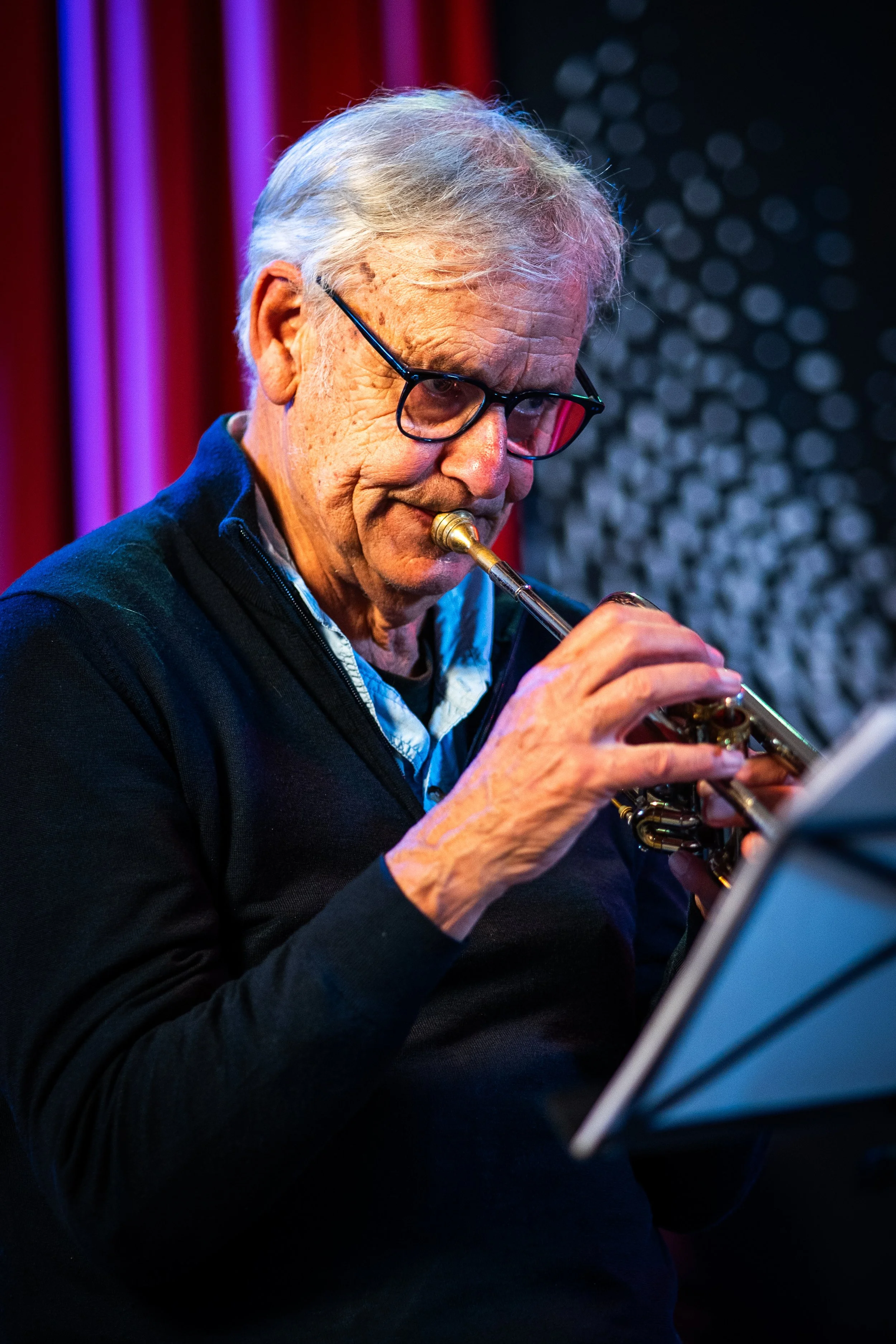 An elderly man wearing glasses plays the trumpet in a dimly lit room with red and purple lighting, and a patterned wall in the background.