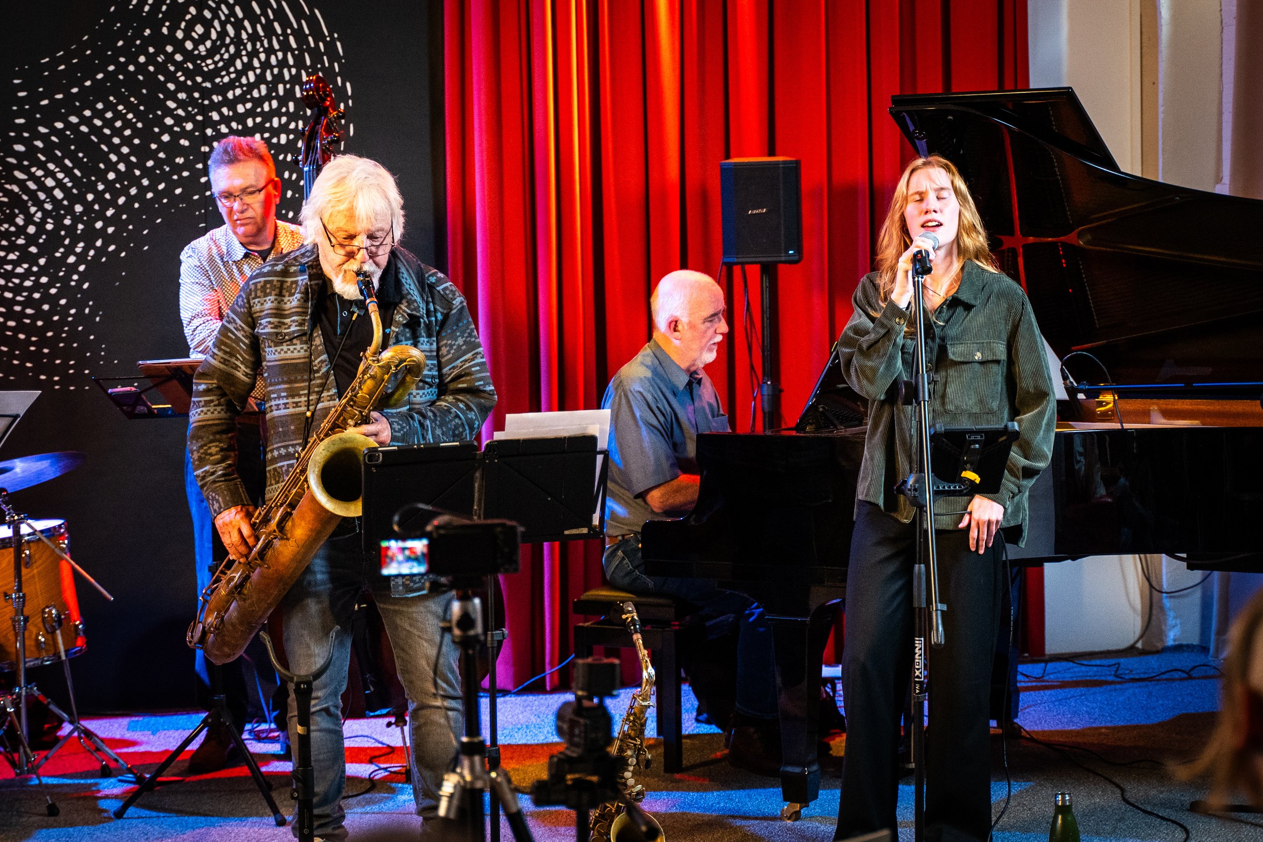 Jazz band performing on stage with four musicians: a saxophonist, a pianist, a double bass player, and a singer, against a backdrop of red curtains and artistic black and white wall design.