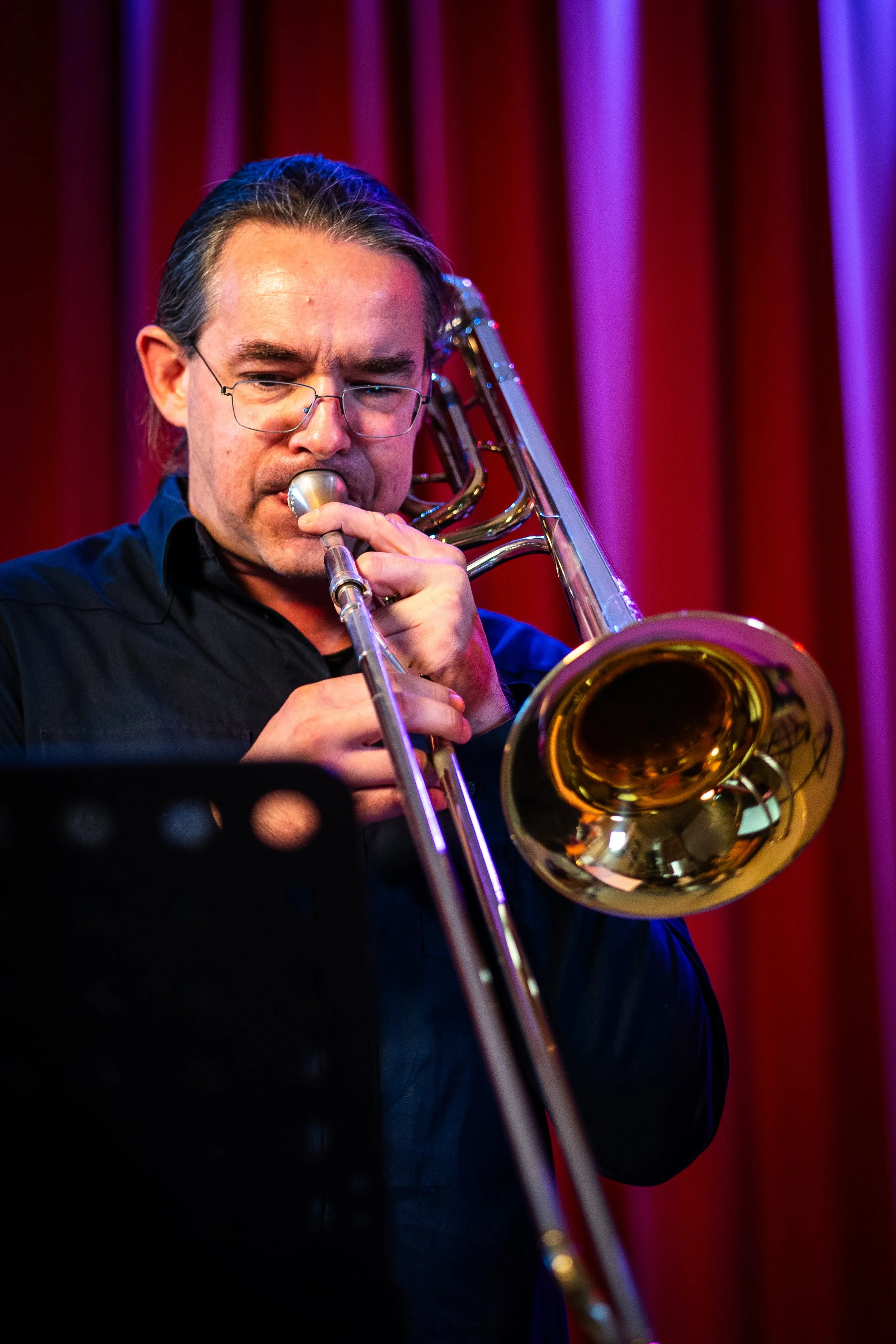 A man playing a trombone on stage with red and purple curtains in the background.