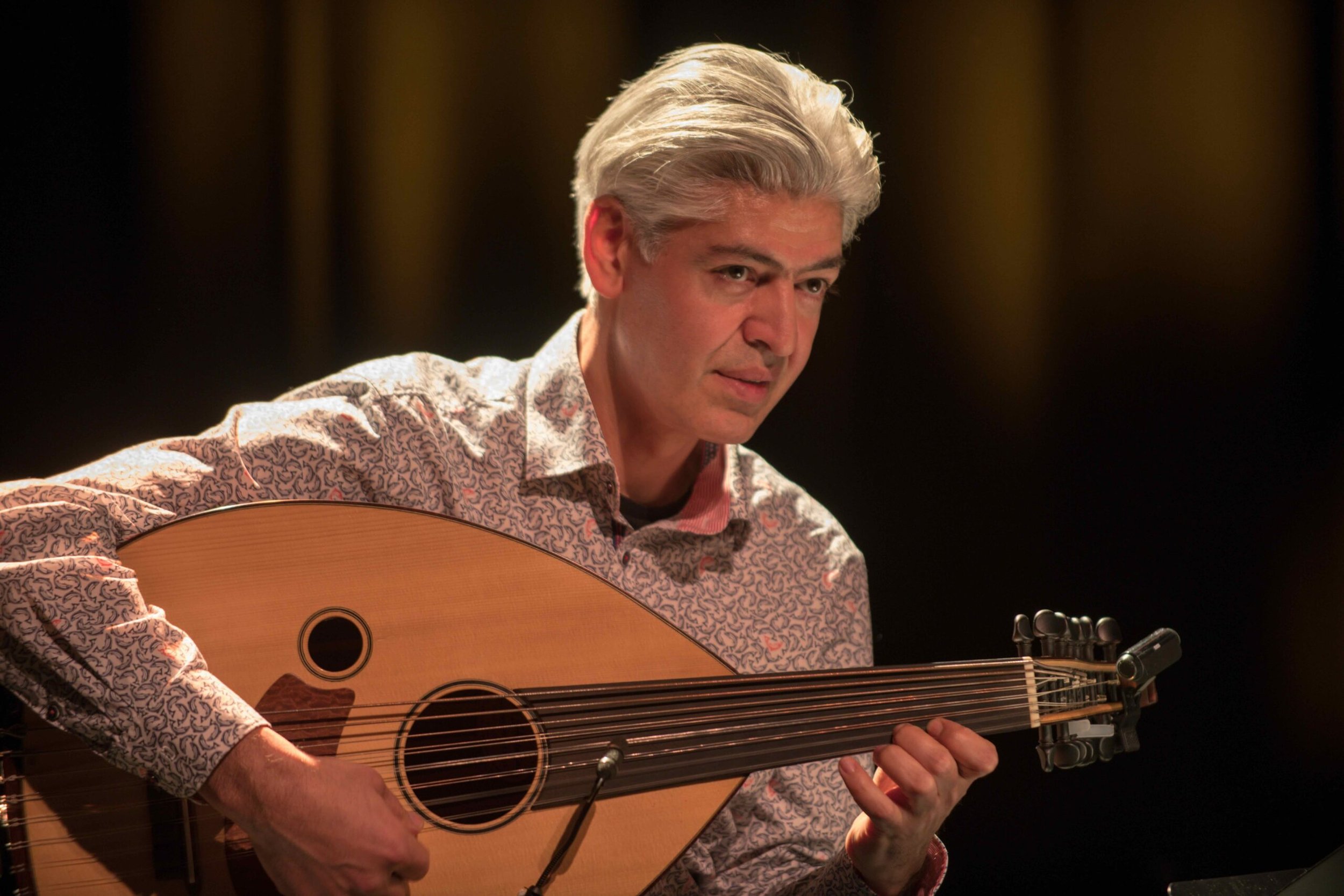 A man with silver hair wearing a patterned shirt is playing an acoustic guitar.