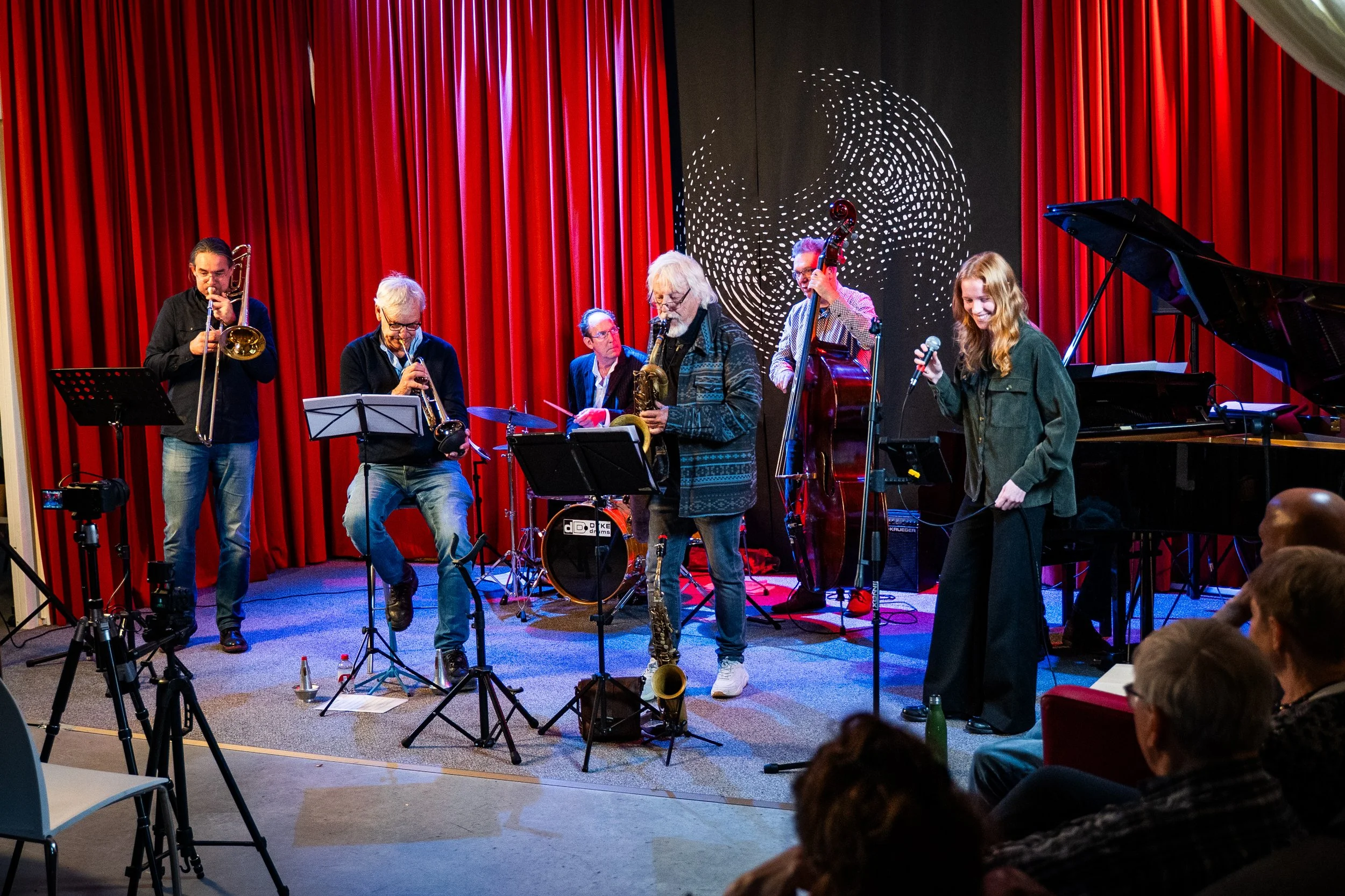 A jazz band performing on stage with red curtains in the background, including musicians playing trumpet, saxophone, double bass, and piano, with an audience watching.