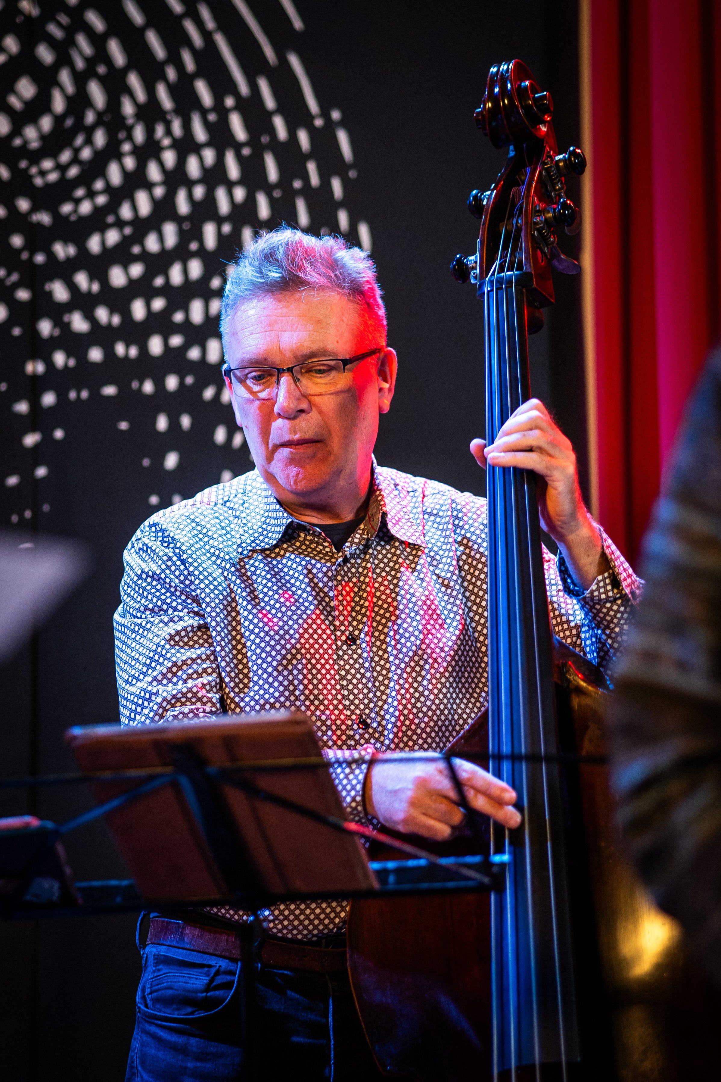 A man with gray hair, glasses, and a patterned shirt playing an upright bass instrument during a performance in a dimly lit setting.