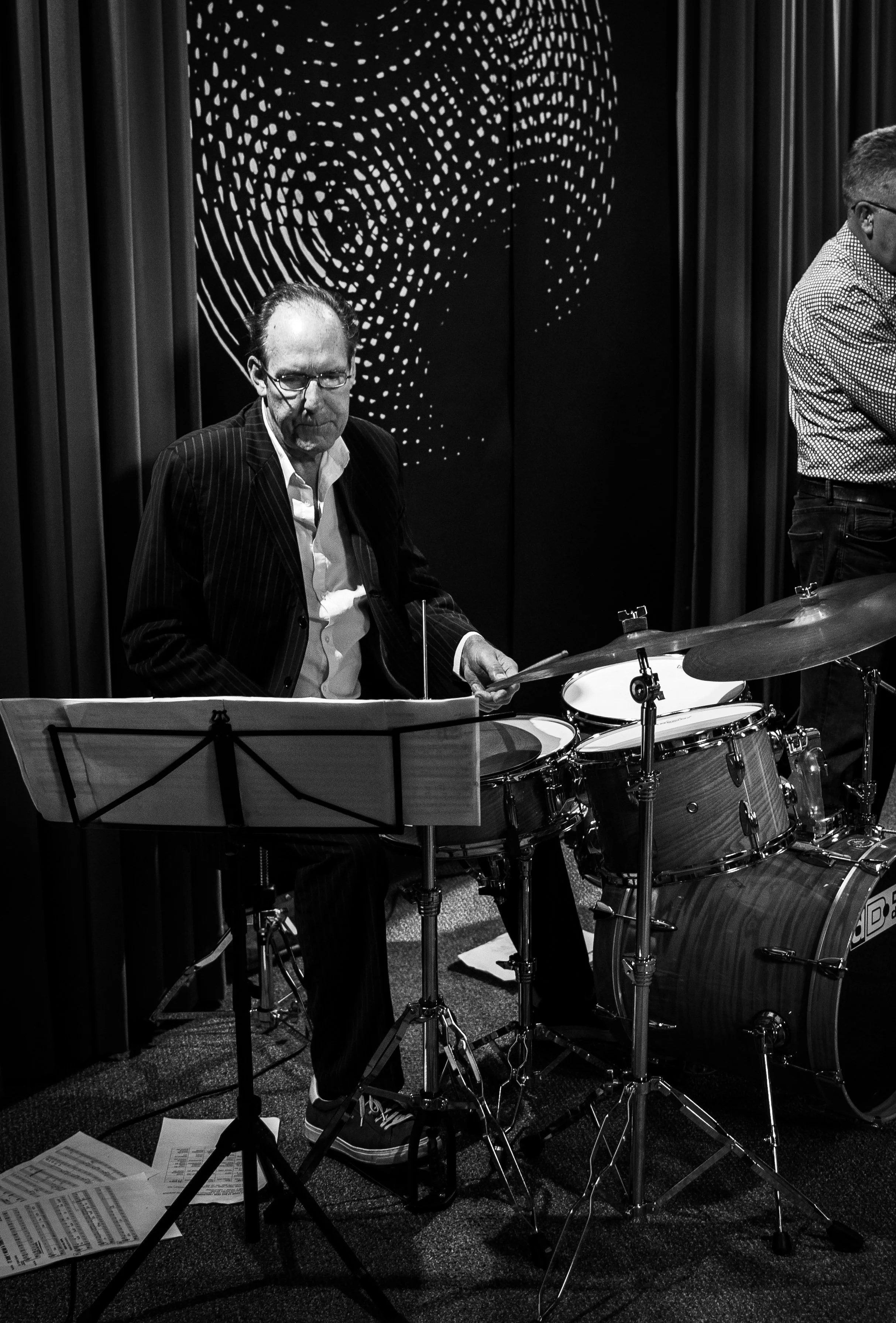 A man with glasses in a pinstripe suit playing drums in a music studio with sheet music on a stand and drawings on the wall.