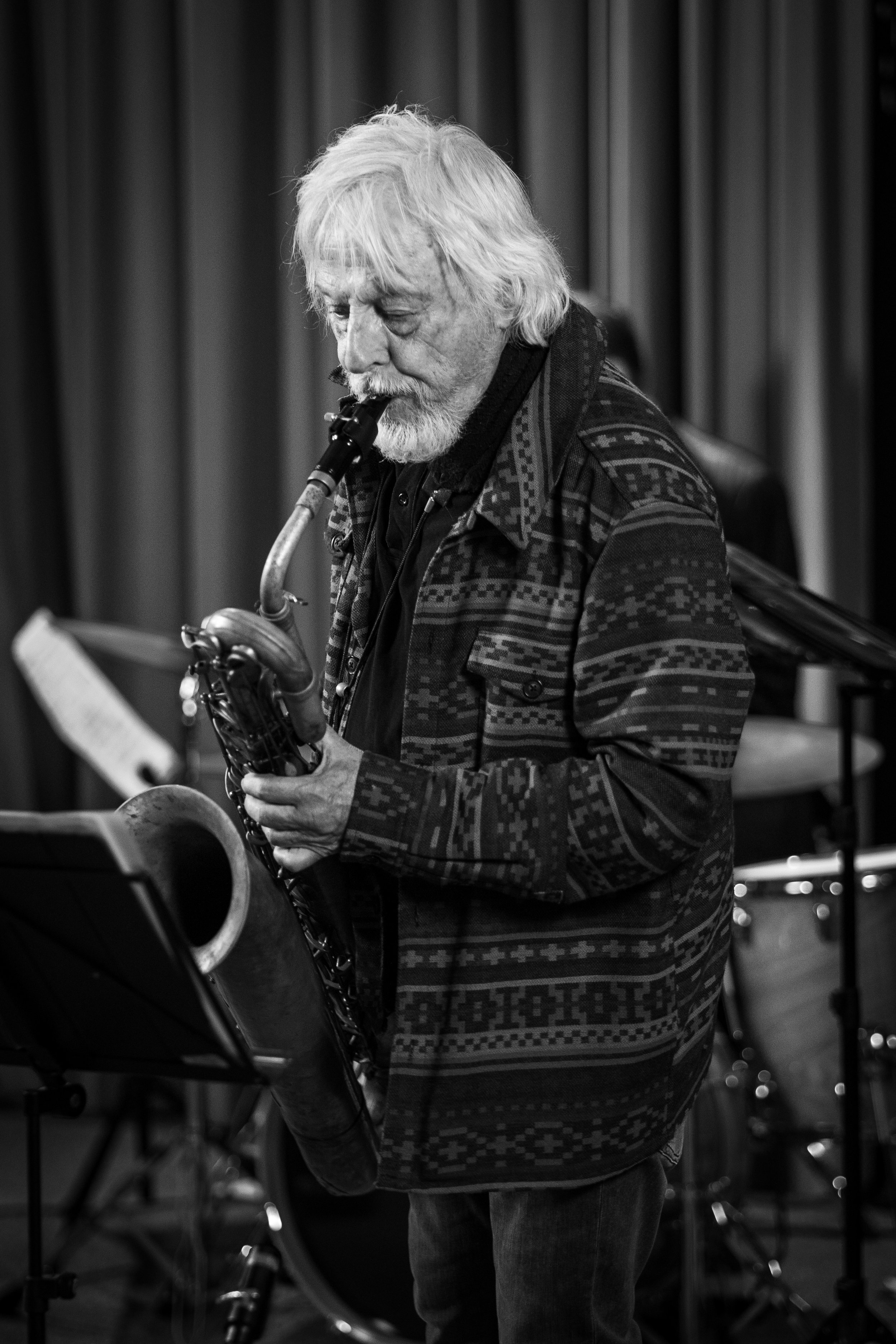 A man with gray hair and a beard playing the saxophone in what appears to be a music studio. He is focused on reading sheet music on a stand and is dressed in a patterned jacket.
