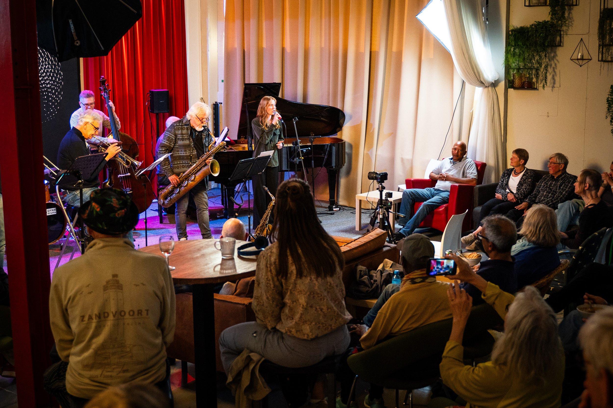 A live jazz band performing on stage with a woman singing into a microphone, surrounded by musicians playing instruments like saxophone, double bass, and piano. Audience seated around the stage, watching and enjoying the performance in a cozy, well-l