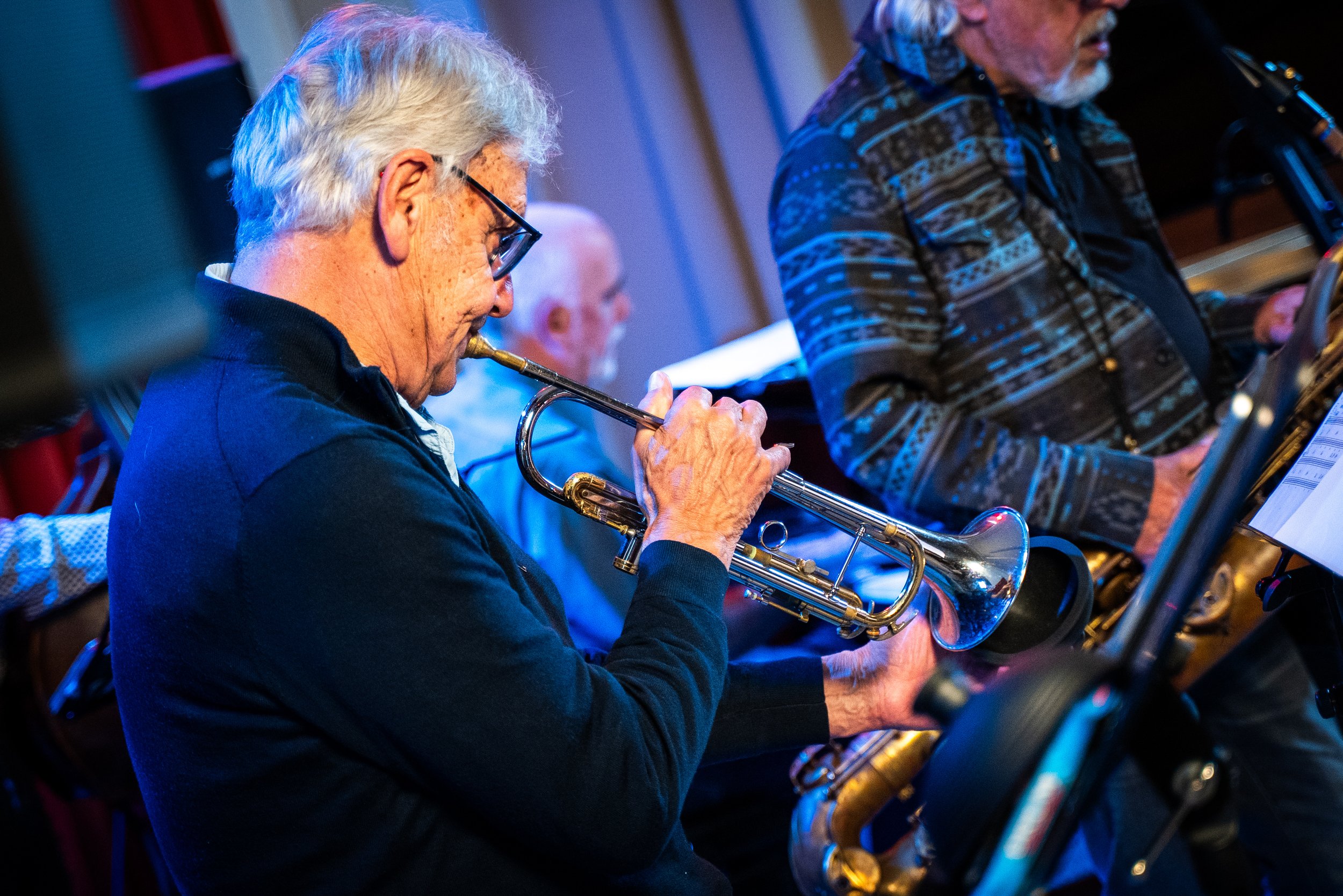 Older man playing trumpet during a band performance, wearing glasses and a dark jacket, with other musicians in the background.