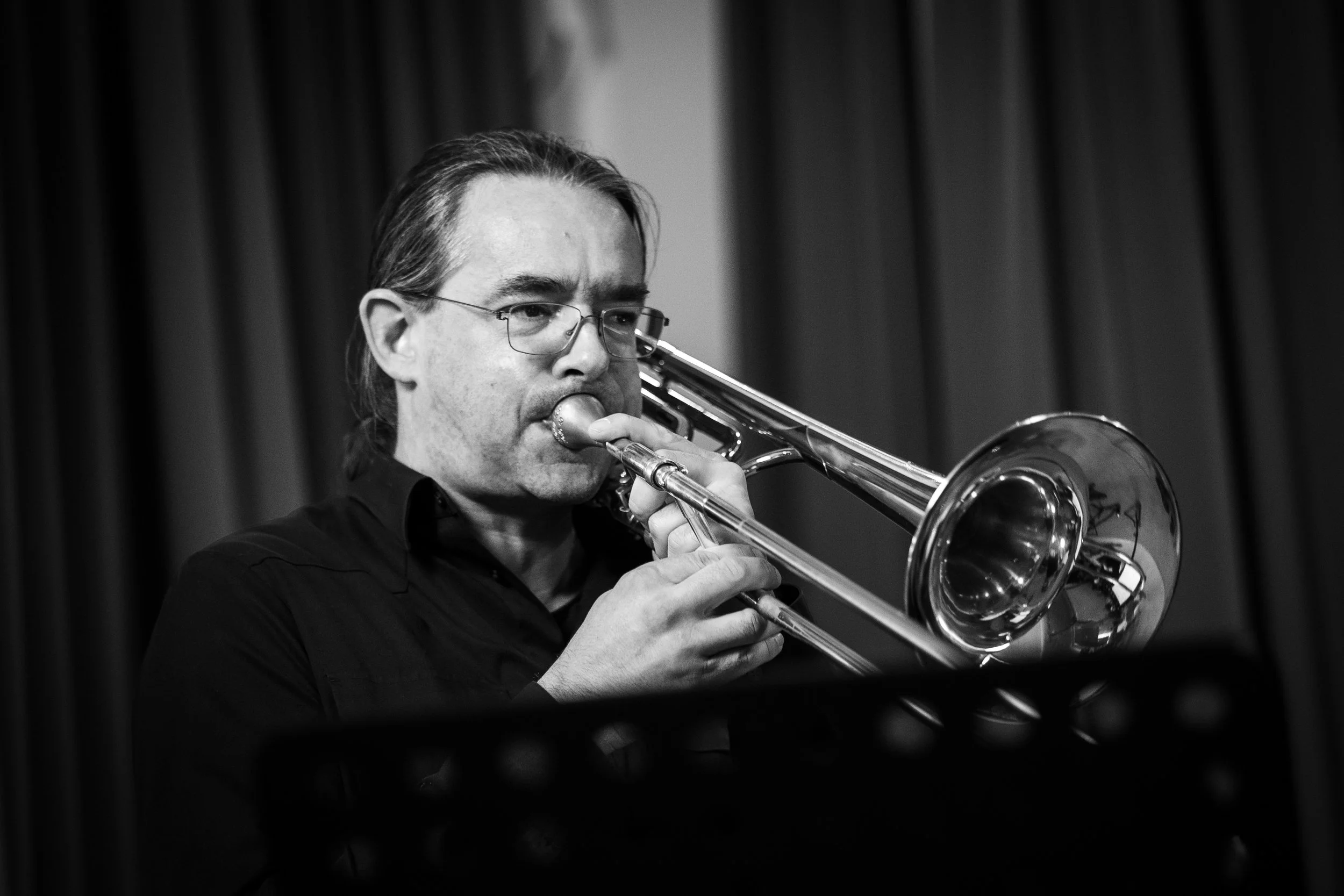A man with glasses playing a trumpet in front of dark curtains.