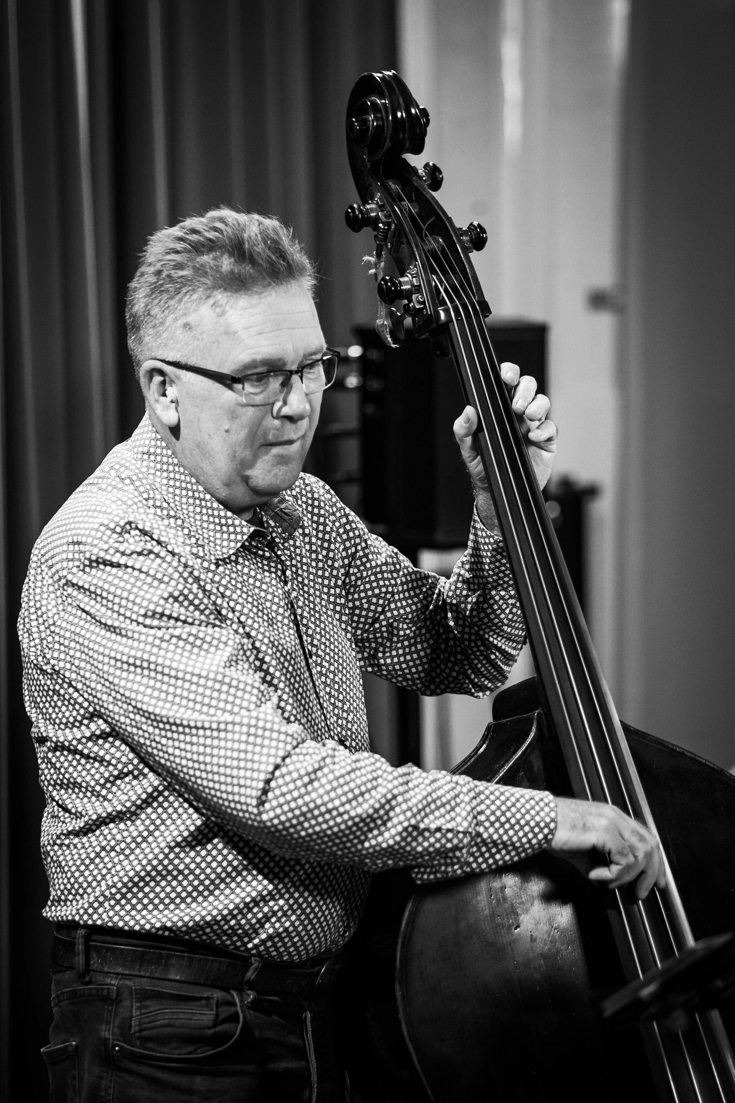 Man playing double bass in a music rehearsal or performance, wearing glasses and a checkered shirt in black and white.