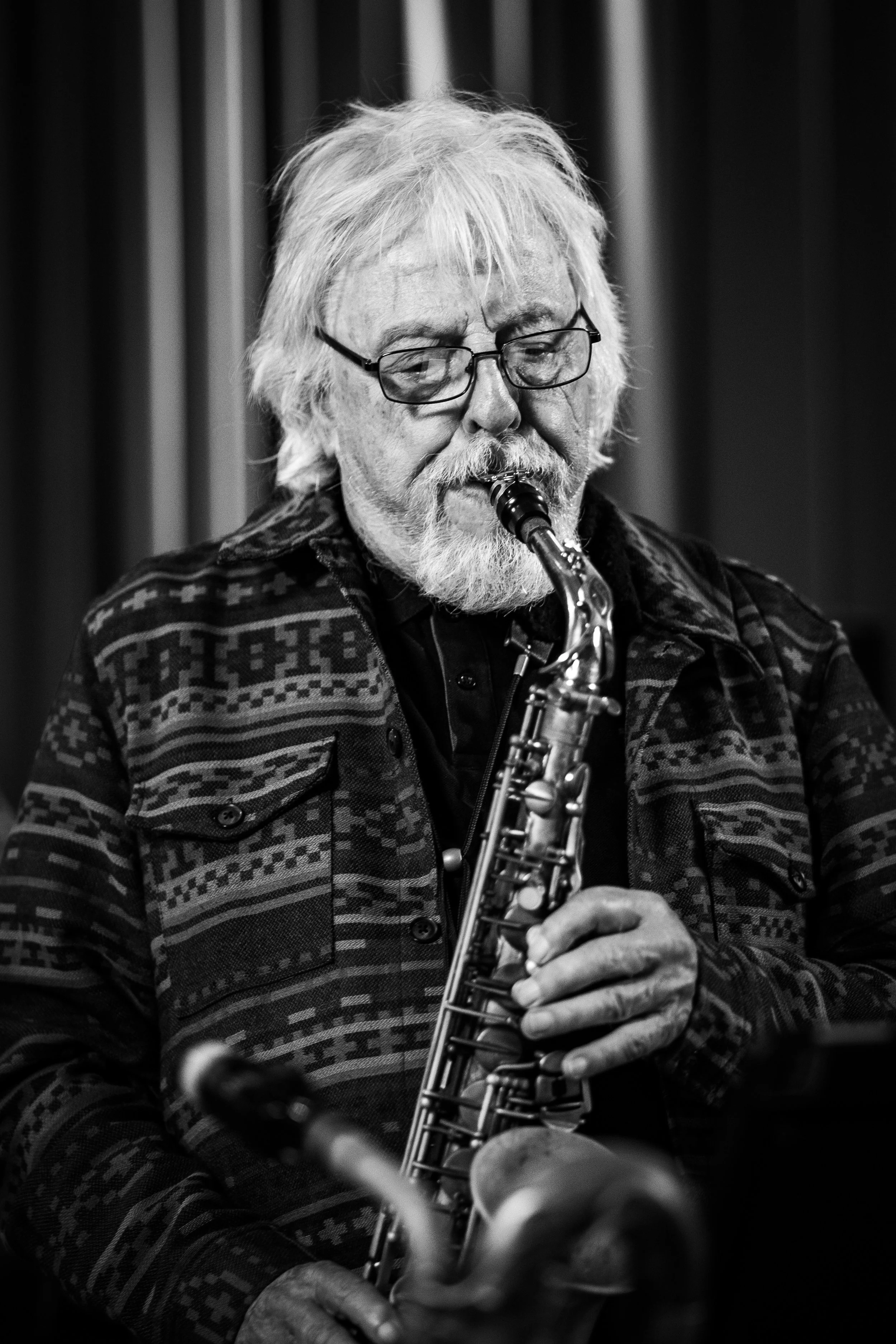 Black and white photo of an elderly man with glasses playing a saxophone, wearing a patterned sweater, in front of a backdrop with vertical curtains.