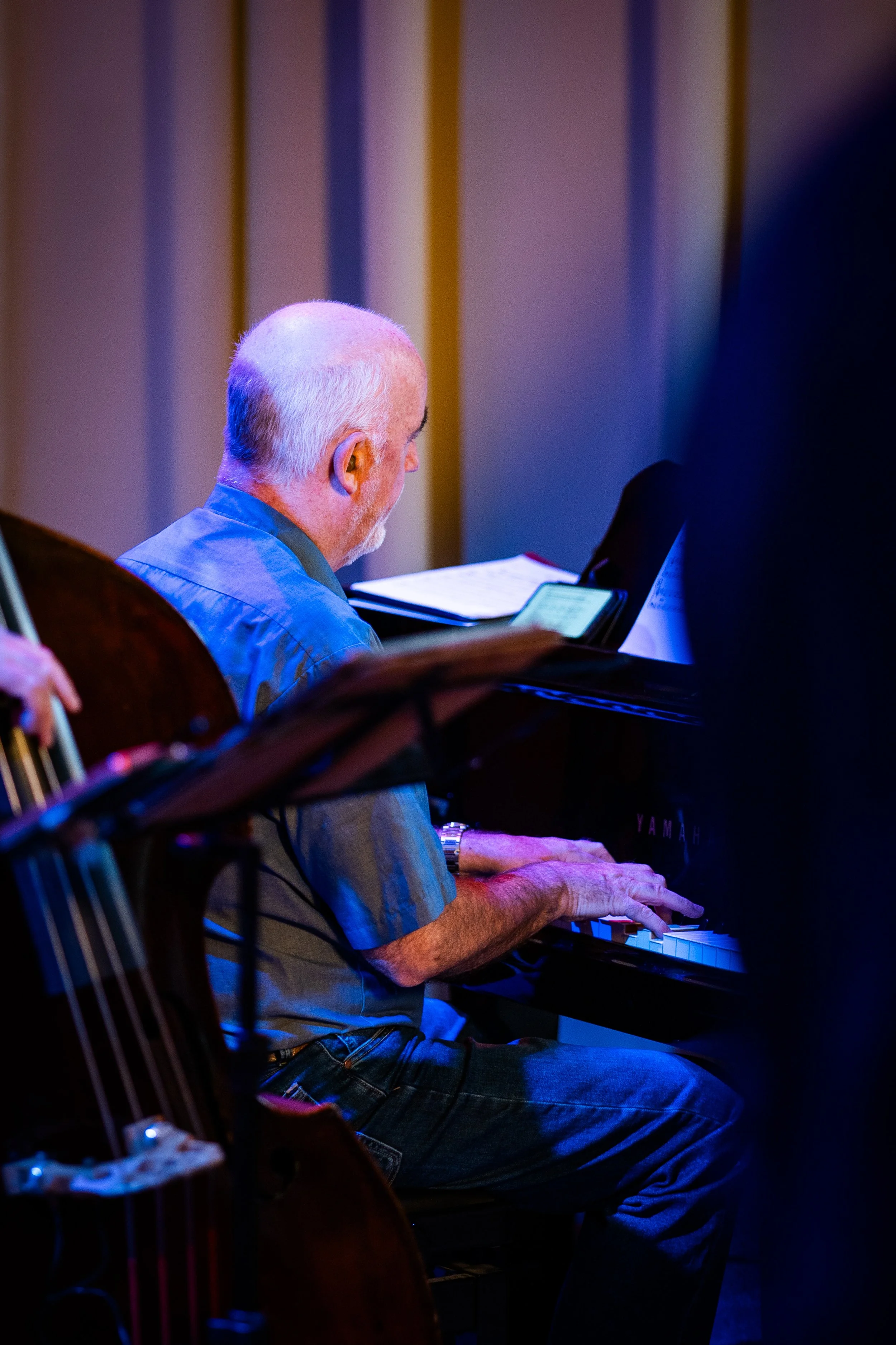 An elderly man with white hair and a beard playing a piano indoors. He is looking at sheets of music and wearing a button-up shirt, illuminated by dim, colorful lighting.