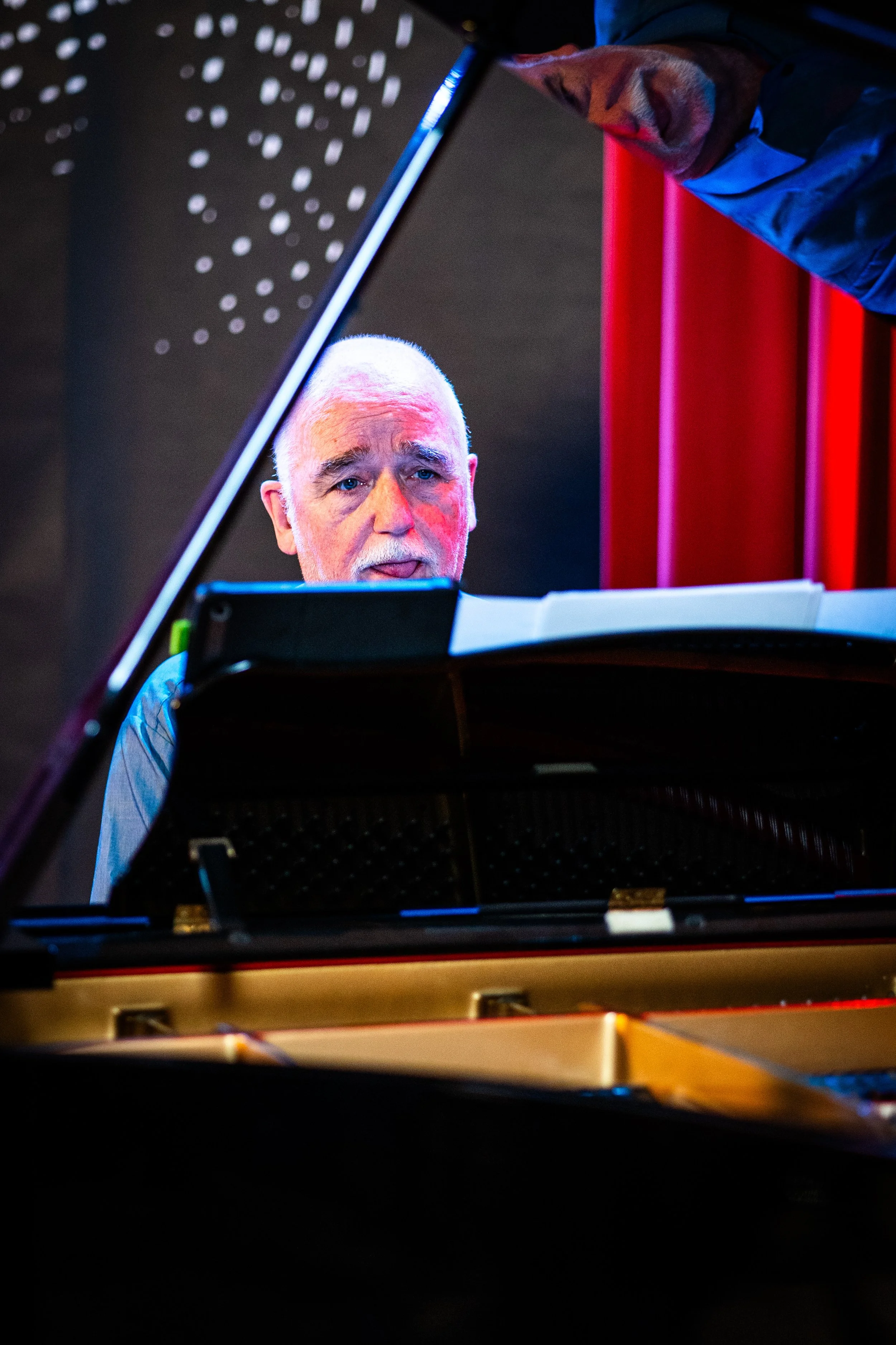 Close-up of an older man with white hair and a beard playing a grand piano with a focused expression.