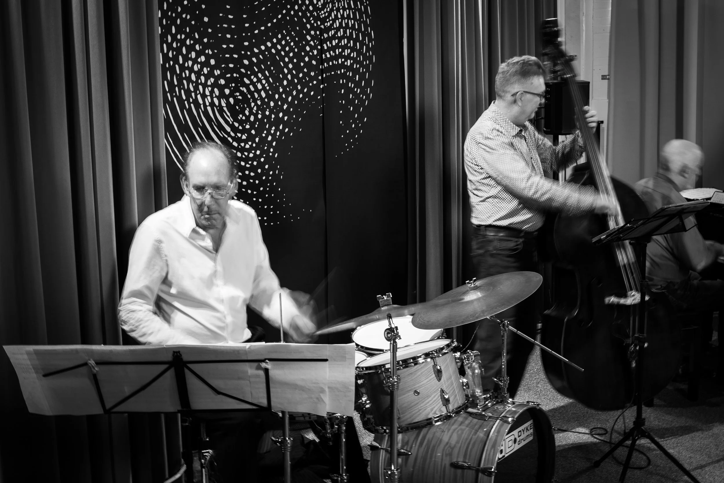 Jazz band performing on stage with a drummer, a bassist, and a pianist, in a venue with dark curtains and a decorative backdrop.