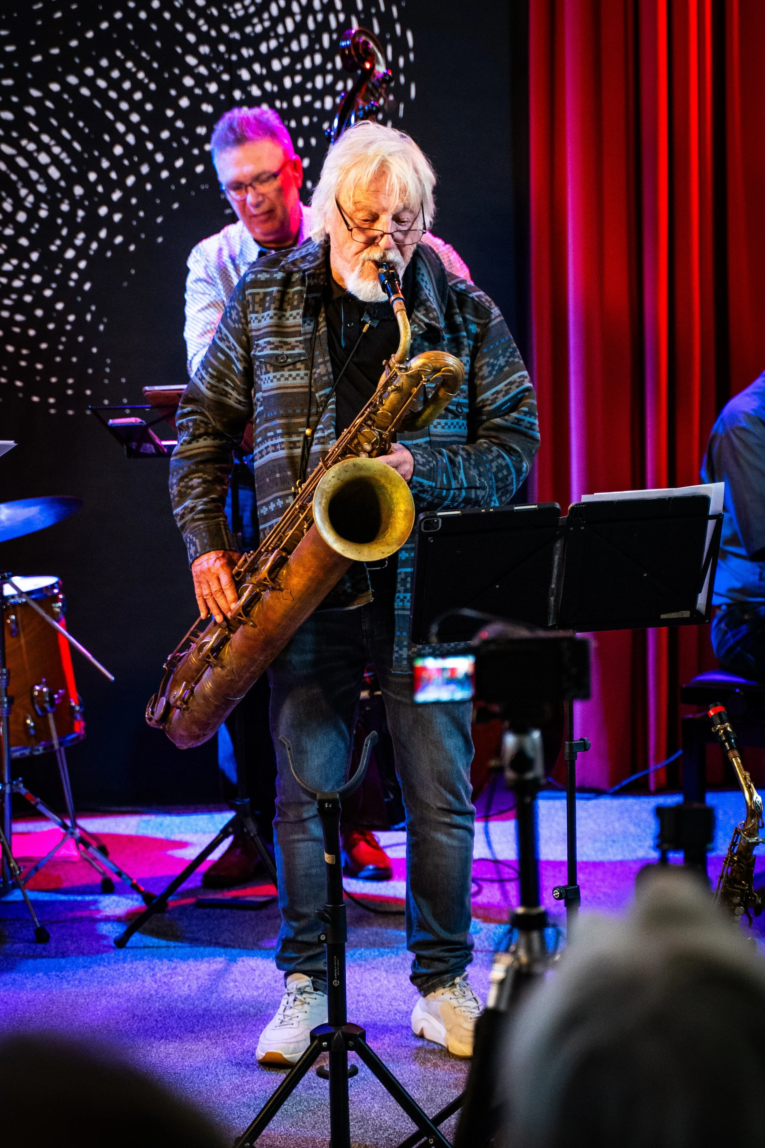 An elderly man with white hair, glasses, and a beard playing a tenor saxophone on stage. He is wearing a black t-shirt, a patterned jacket, jeans, and white sneakers. Behind him, a man with glasses and gray hair is playing a bass guitar. The stage ha