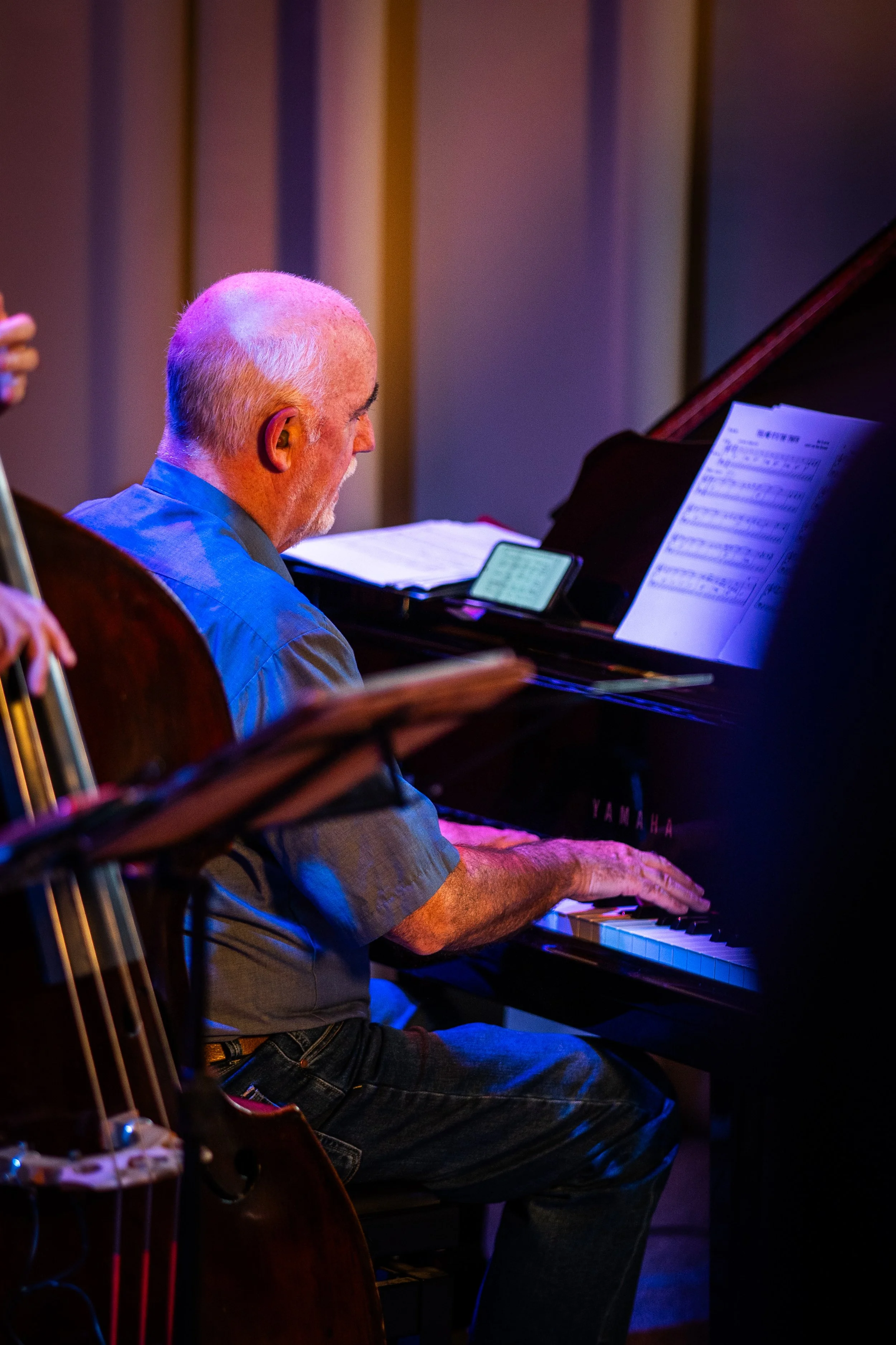 An older man playing the piano, wearing a gray shirt, with sheet music on the piano and a mobile phone on a stand nearby, in a dimly lit room with beige vertical wall panels.