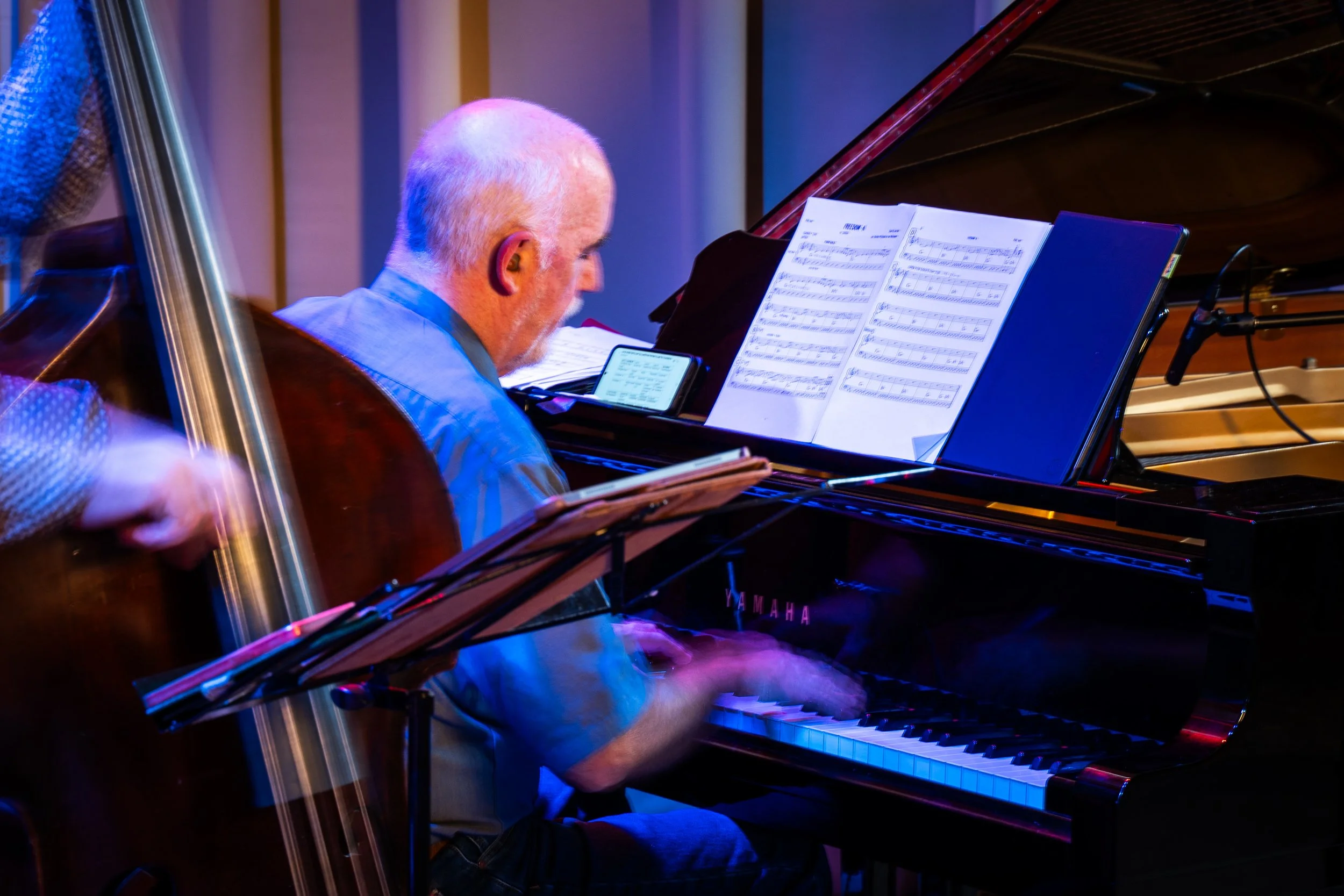 An older man with short white hair is playing a grand piano, reading sheet music, in a performance setting with colorful lighting.