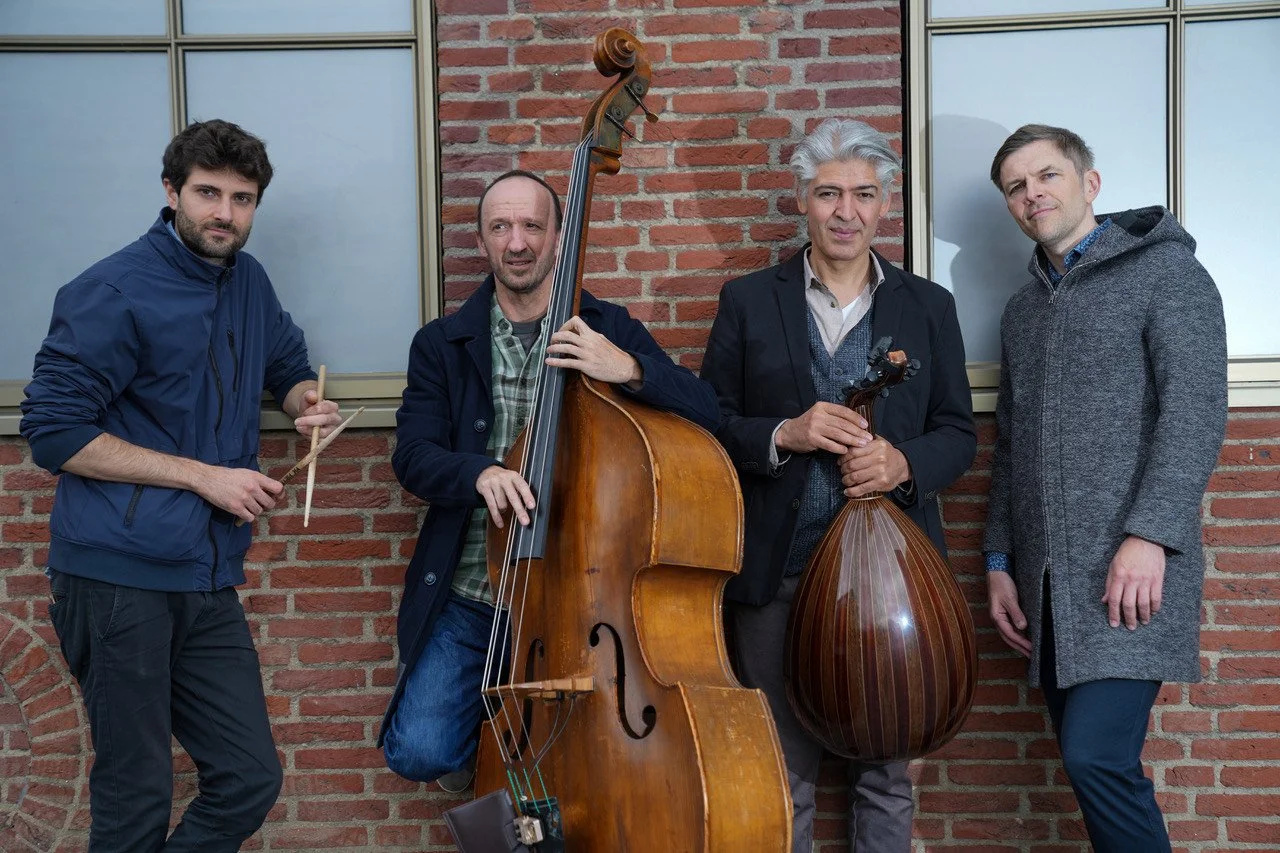 Four men standing against a brick wall, with musical instruments including a drum, double bass, and sarang while looking at the camera.