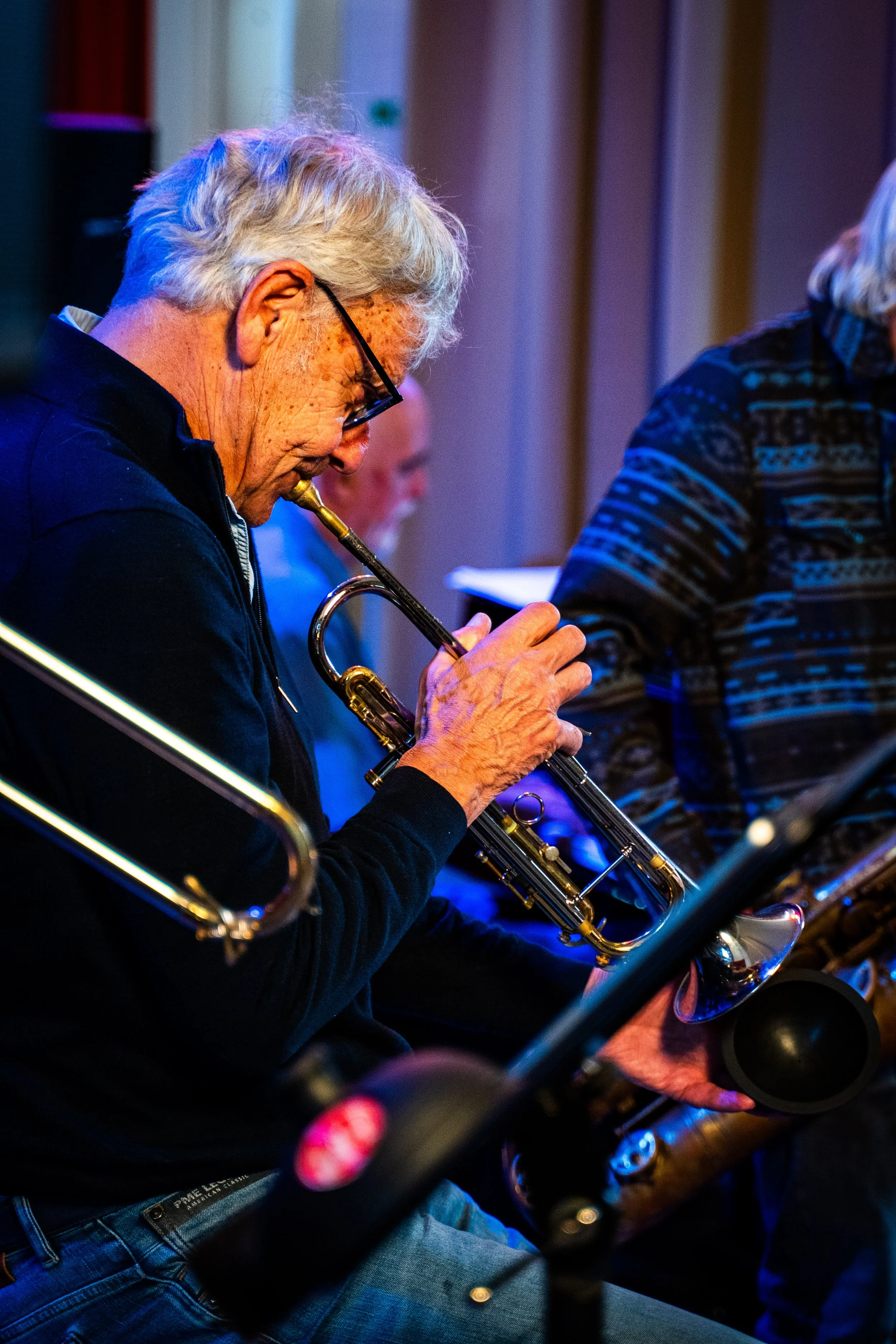 An elderly man with gray hair, glasses, and freckles playing a trumpet. He is seated and focused on his music in a dimly lit setting, possibly during a live performance or practice session.