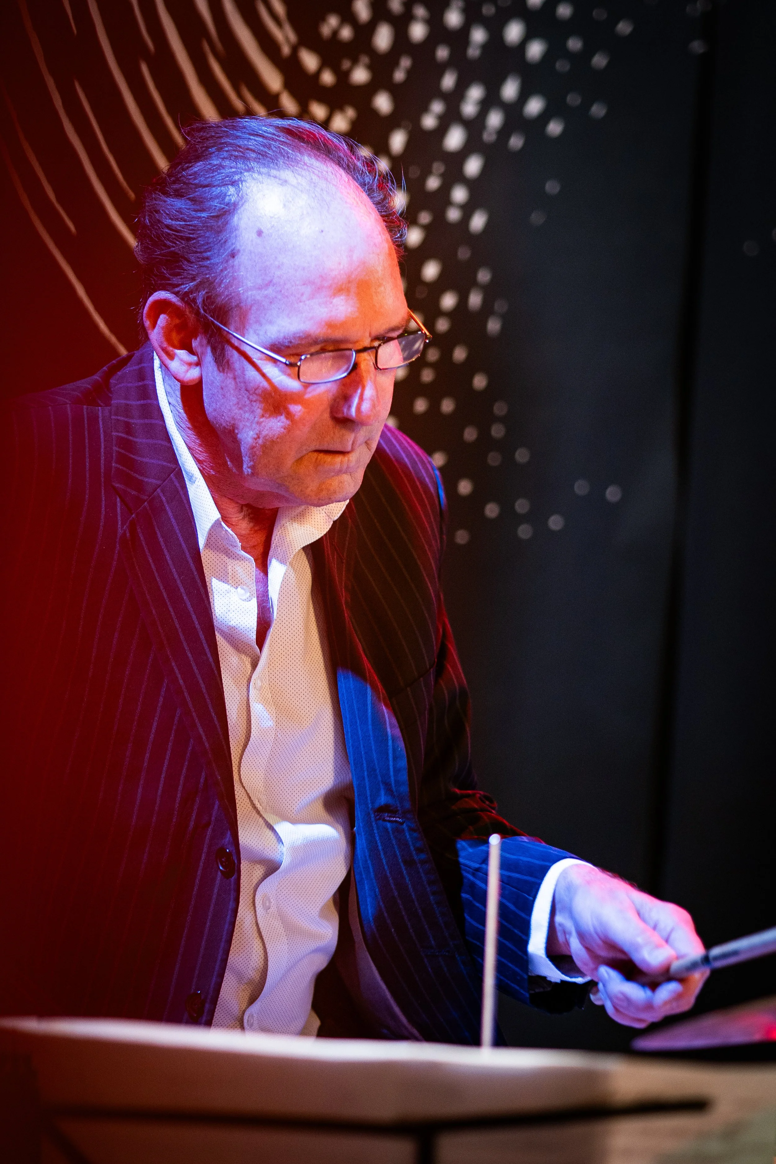 A man with glasses in a pinstripe suit is looking down at a device, likely a tablet, in a dimly lit environment with colorful lighting. Abstract geometric patterns are visible in the background.