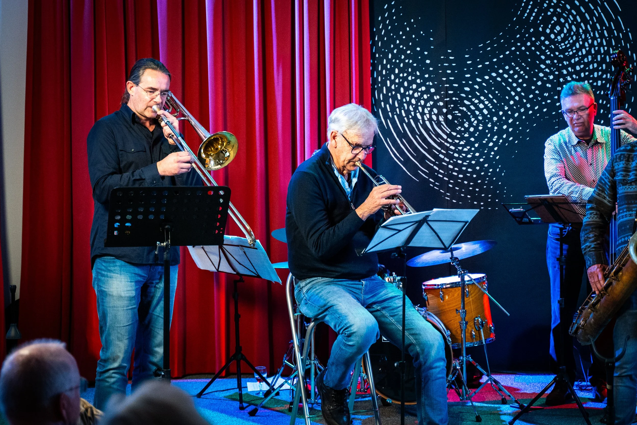 Three musicians performing on stage with a red curtain and a digital background, one playing trumpet, one playing drums, and another playing a double bass.
