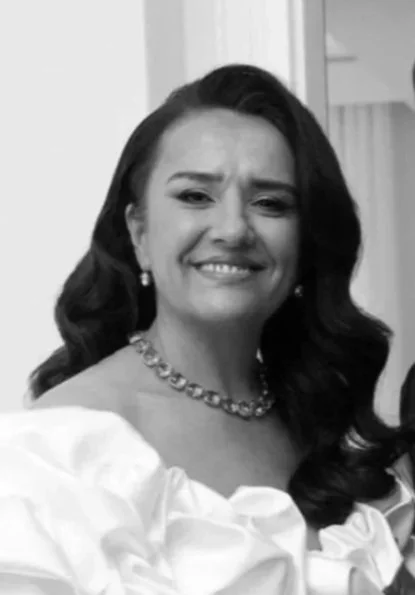 A woman smiling, wearing earrings, a necklace, and a white ruffled dress.