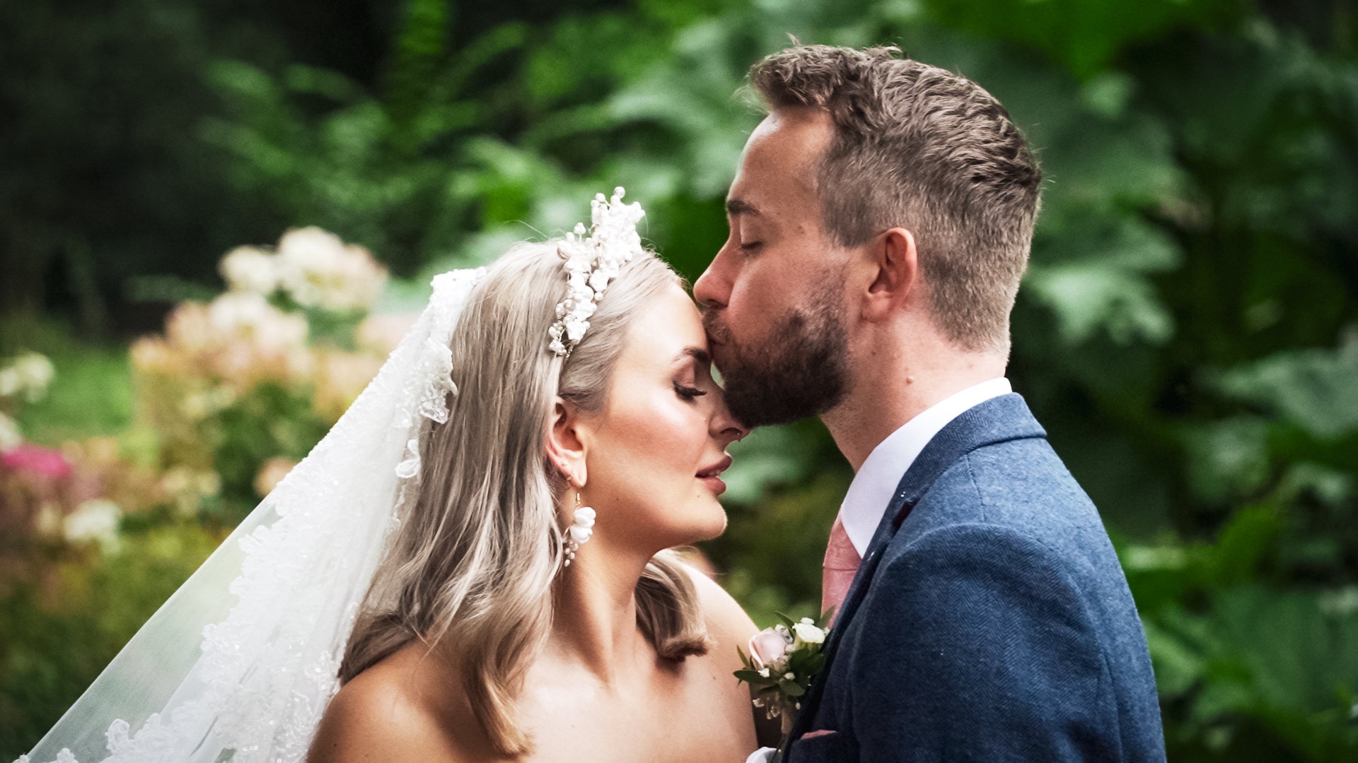 A bride and groom sharing a moment with foreheads touching and eyes closed, outdoors with lush green background.