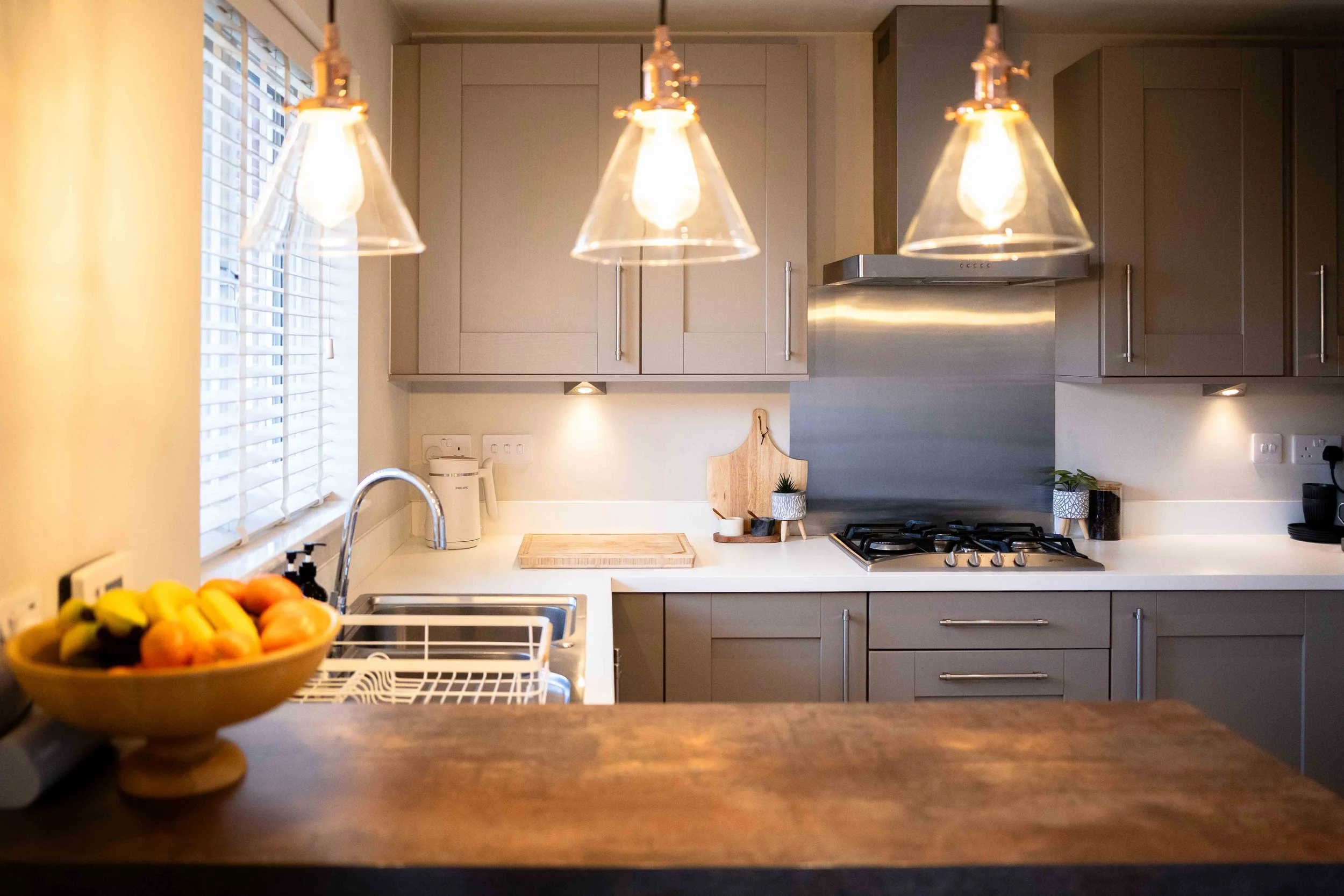 Modern kitchen with grey cabinets, stainless steel range hood, four glass pendant lights, white countertops, and a window with white blinds.