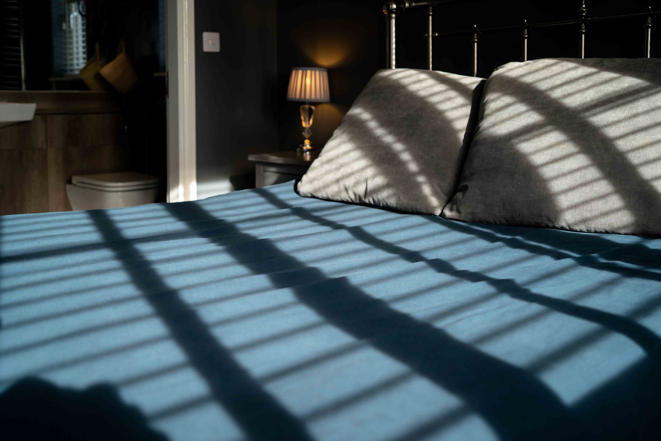 A bed with a blue bedsheet and two gray pillows, shadows from window blinds cast across the bed, and a nightstand with a lamp in a dimly lit bedroom.