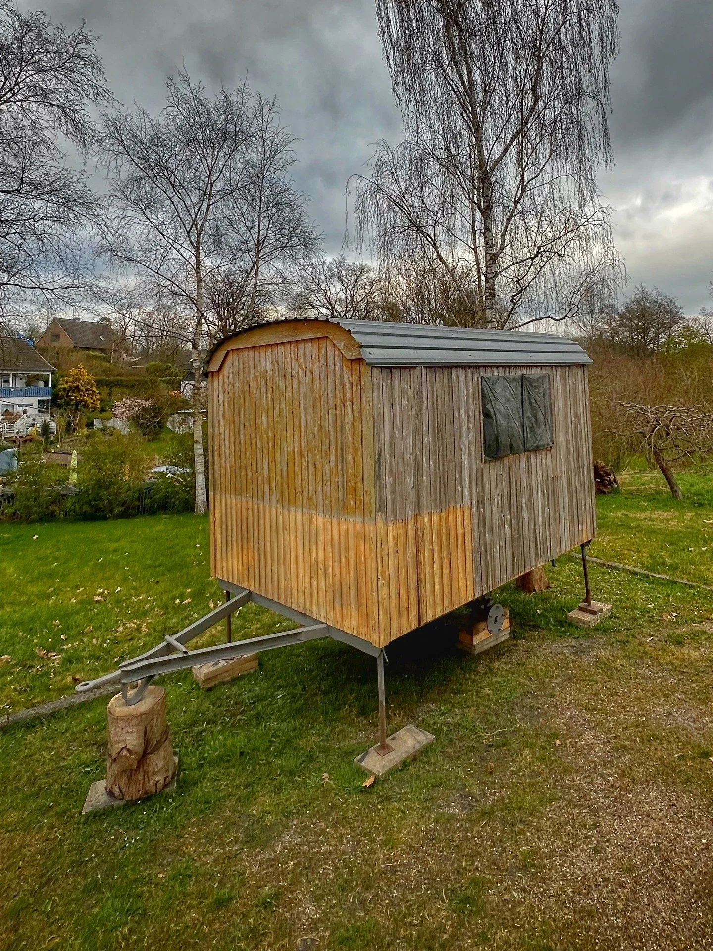 Ein kleiner hölzerner Bauwagen mit Dach und Fenster auf einer Wiese, umgeben von Bäumen und Häusern im Hintergrund unter einem bewölkten Himmel.