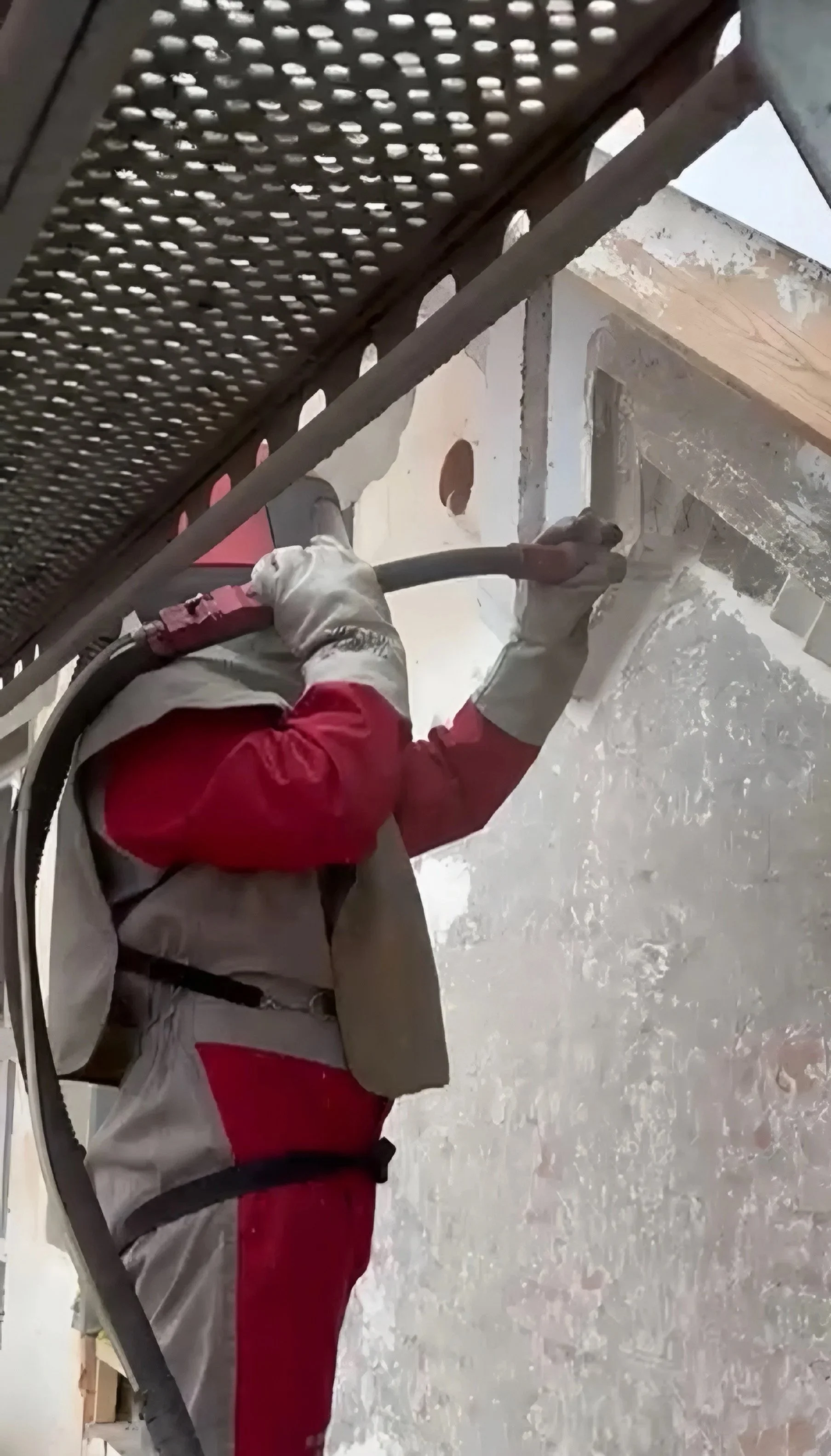 Ein Arbeiter in Schutzkleidung und Handschuhen arbeitet an einer Wand und strahlt diese mit Sand im Aussenbereich eines Gebäudes.