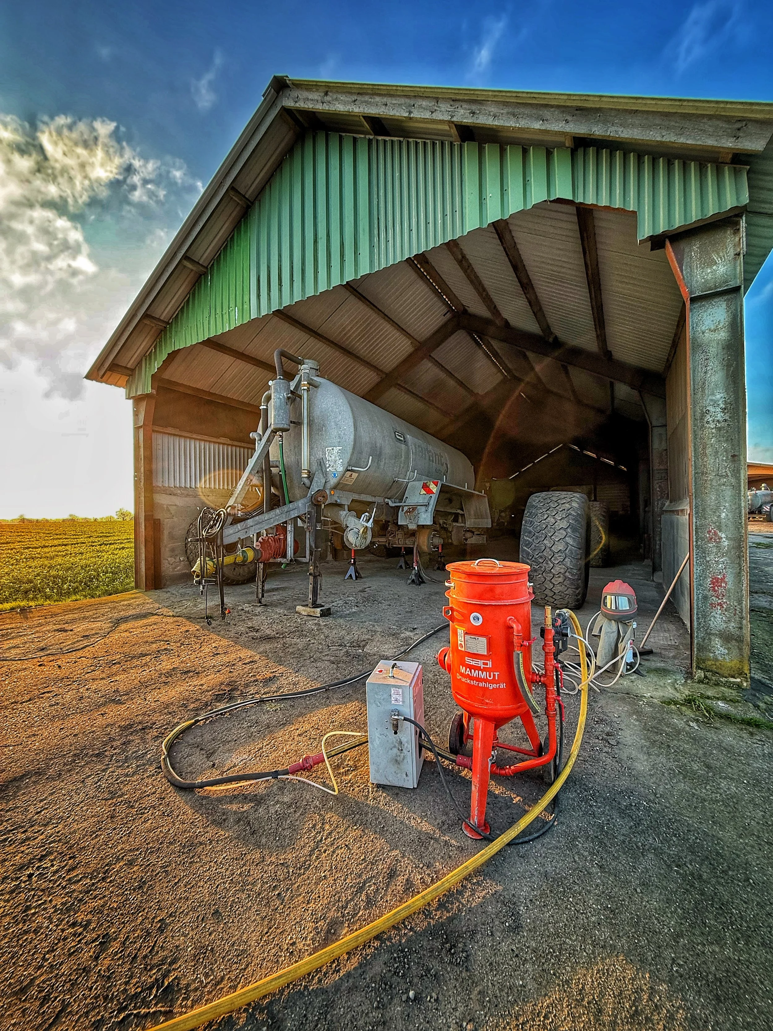 Landwirtschaftlicher Schuppen mit landwirtschaftlichen Geräten und einem Tankwagen, unter blauem Himmel mit Wolken.