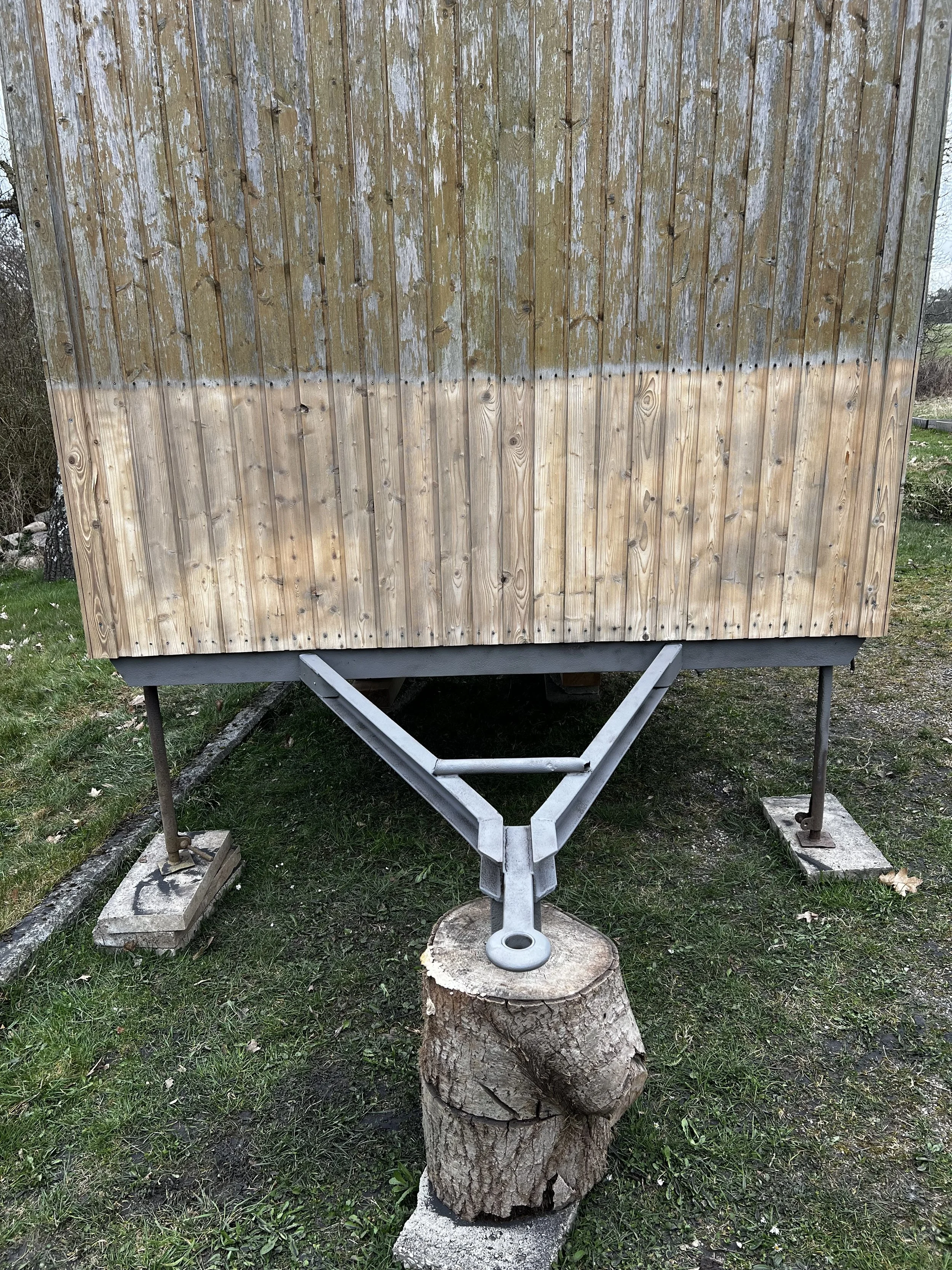 Ein umgebauter Anhänger, dessen Unterseite mit unbehandeltem Holz verkleidet ist, steht auf einem Baumstamm aufgestützt im Freien.