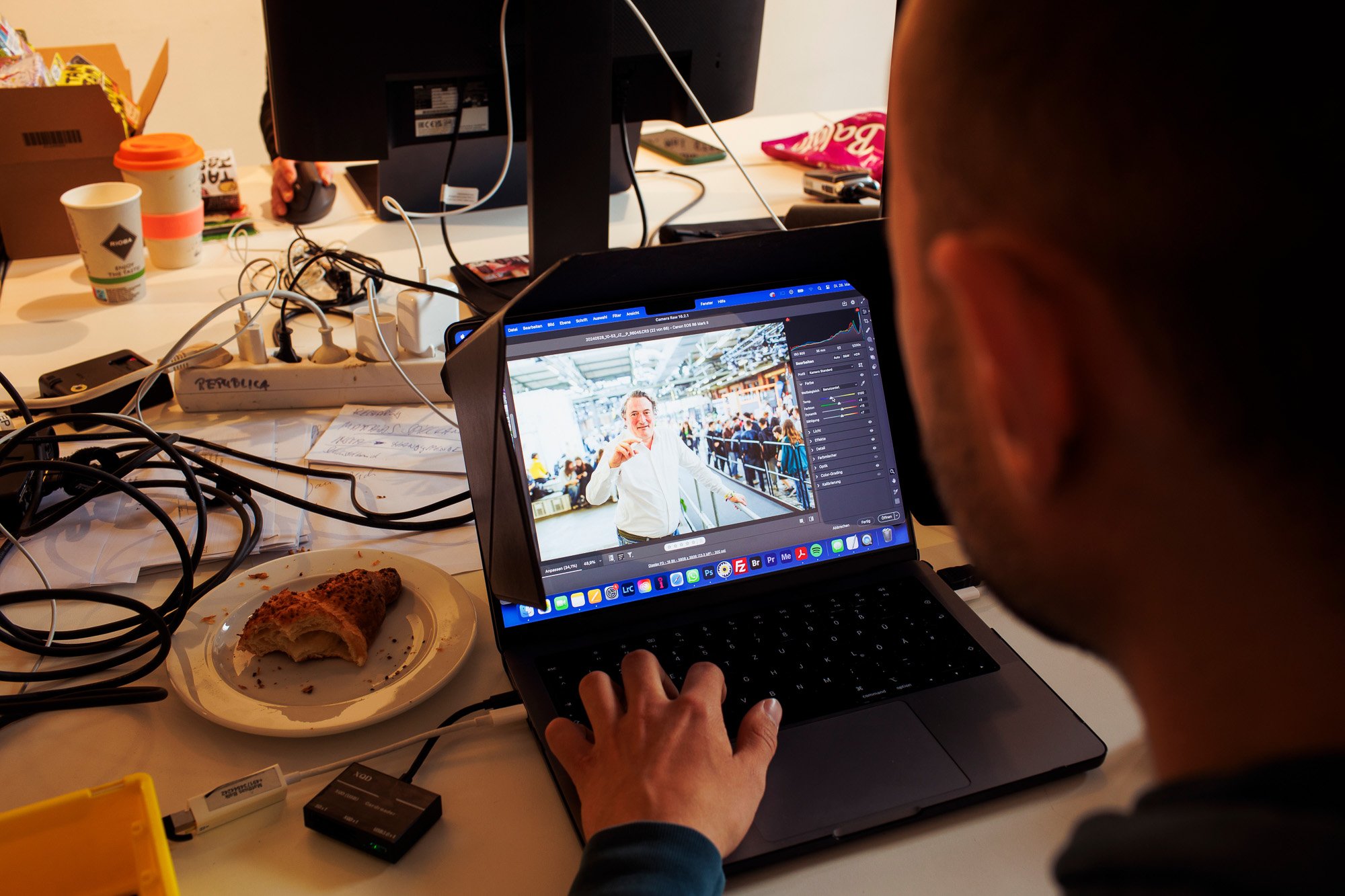 Person am Laptop in einem Büro mit mehreren Bildschirmen und unordentlichem Schreibtisch.