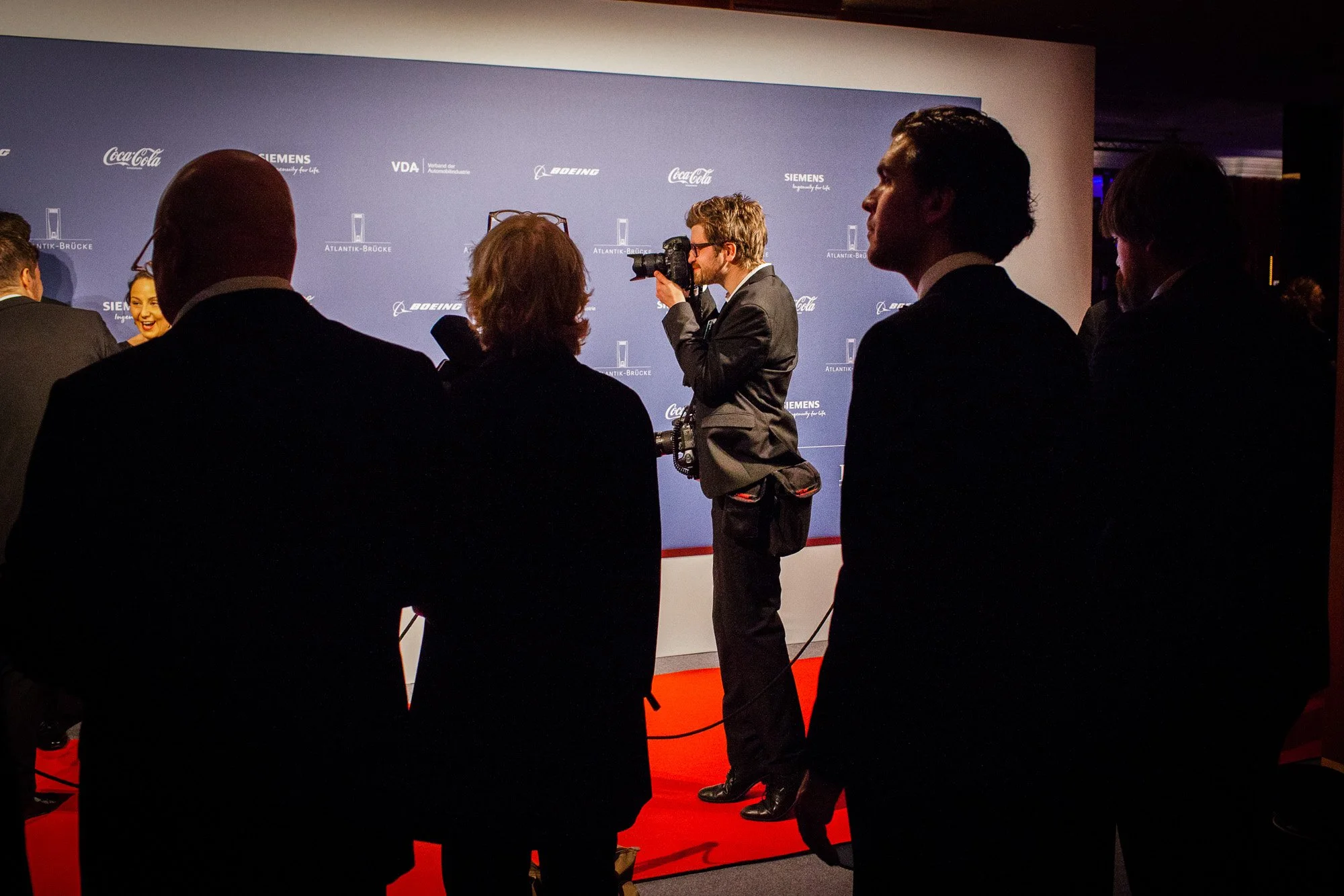Ein Fotograf steht auf einem roten Teppich und macht Fotos von Personen vor einer Sponsorenwand. Die Umgebung ist dunkel und es sind weitere Menschen anwesend.