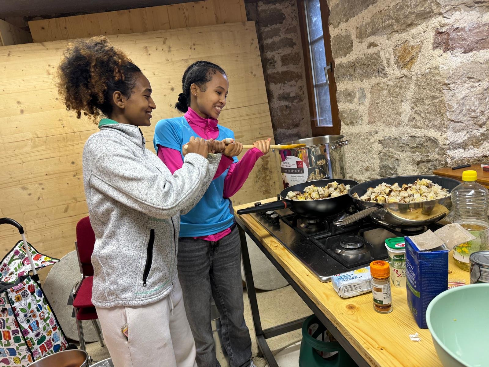 Zwei Frauen kochen gemeinsam in einer Küche. Sie stehen nebeneinander an einem Herd, auf dem Pfannen mit Essen stehen. Die Umgebung sieht rustikal mit einem Steinwandhintergrund aus. Auf dem Tisch liegen verschiedene Kochutensilien und Zutaten.