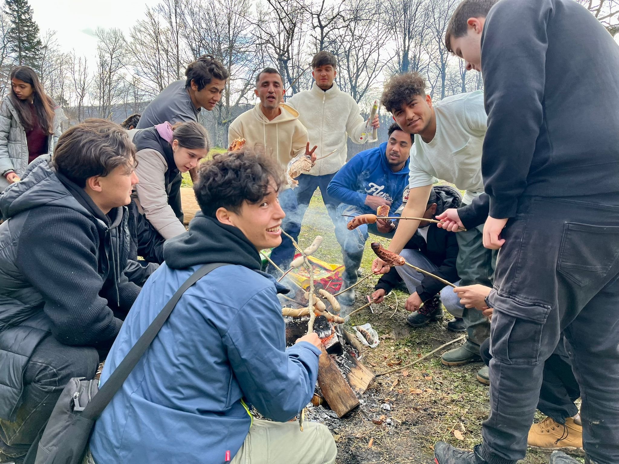 Eine Gruppe von Menschen grillt Würste über einem Lagerfeuer im Freien. Sie lachen und genießen die gemeinsame Zeit.