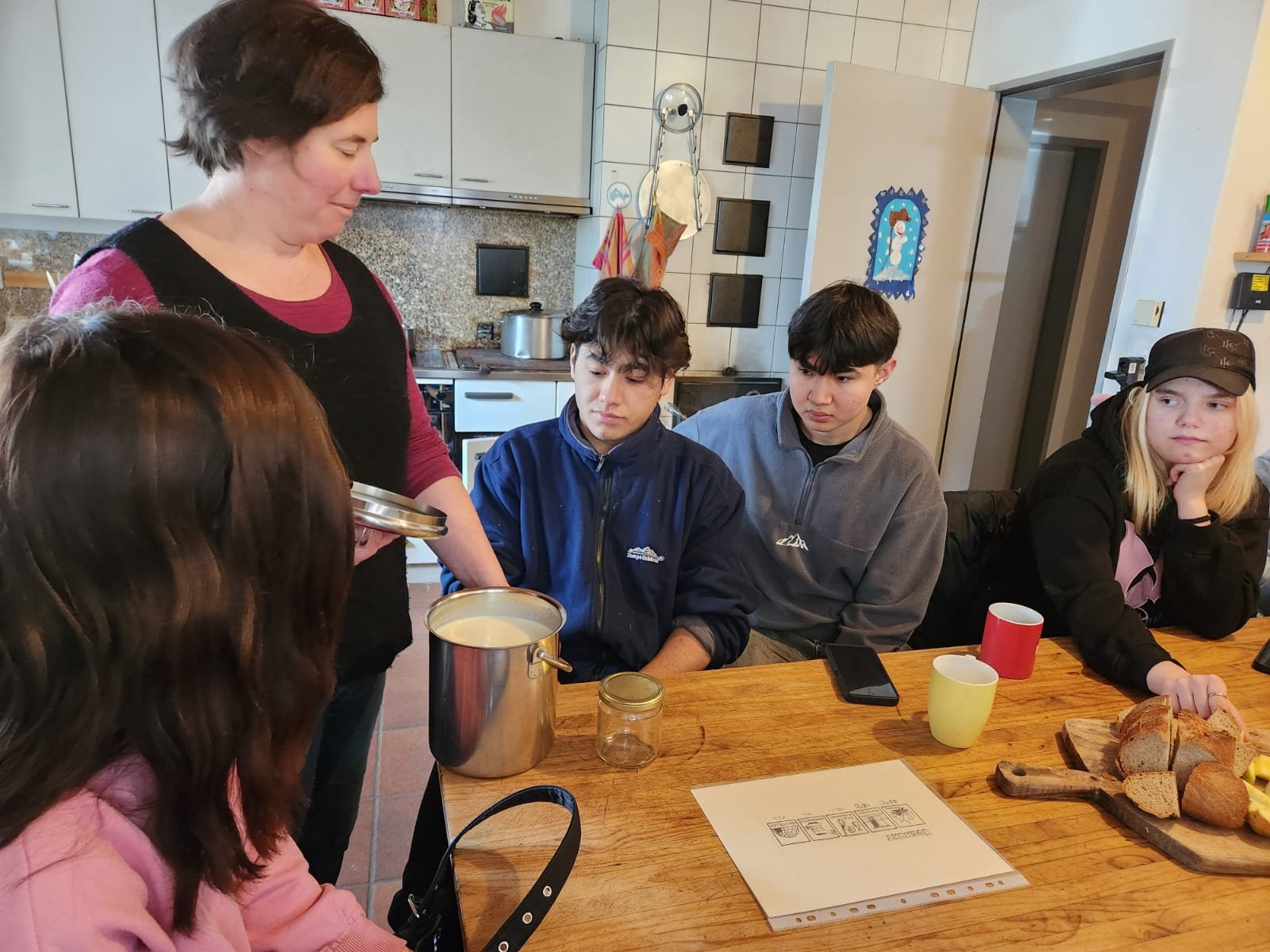 Eine Frau zeigt einer Gruppe von jungen Menschen einen Topf mit Milch. Sie sitzen in einer Küche an einem Tisch mit Bechern und Brotscheiben auf einem Holzbrett. Auf dem Tisch liegt außerdem ein Blatt Papier.