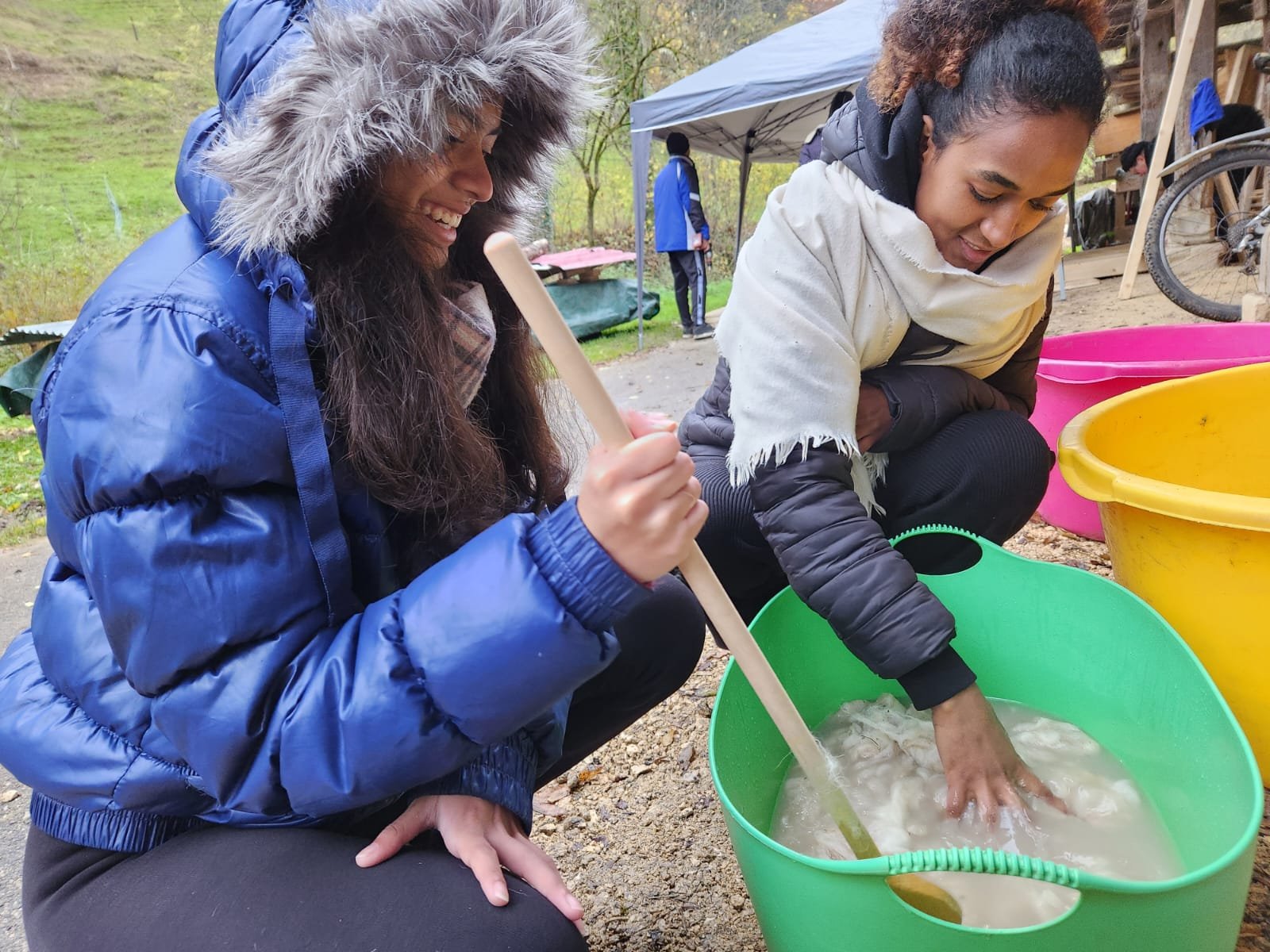 Zwei Menschen interagieren mit einer großen grünen Wanne, die mit Wasser und einer weißen Substanz gefüllt ist. Eine Person rührt mit einem Stock um, während die andere ihre Hand im Wasser hat. Beide tragen warme Kleidung und die Szene findet im Frei