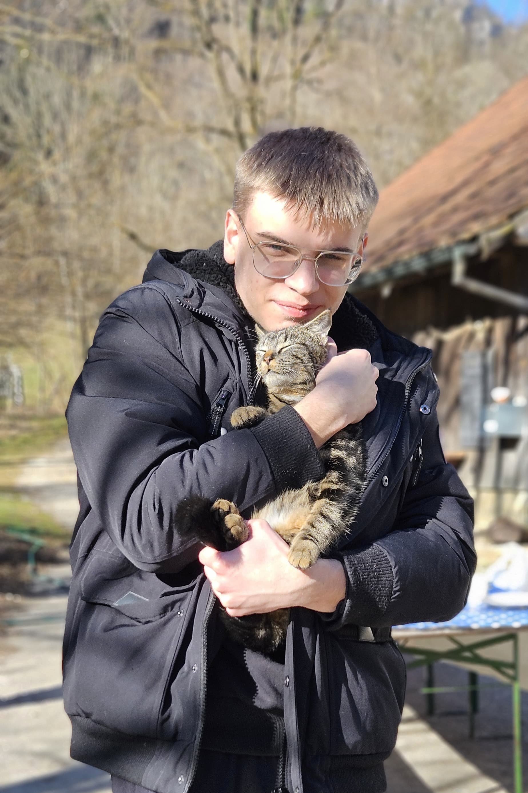 Eine Person in einer schwarzen Jacke hält eine getigerte Katze im Arm, im Hintergrund ein Gebäude mit Holzdach und Bäume.