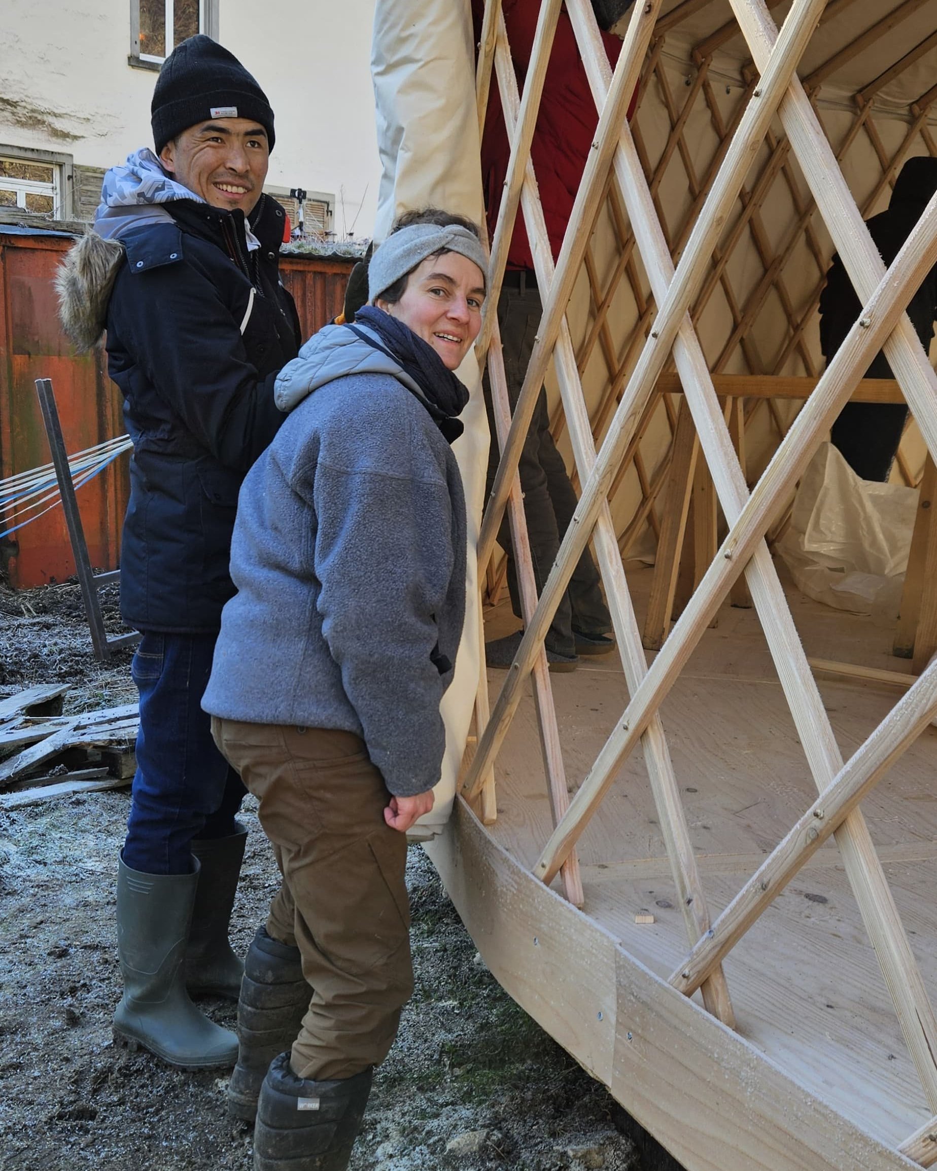 Zwei Personen bauen eine Holzstruktur auf, die wahrscheinlich Teil eines Jurten-Zelts ist. Beide tragen warme Kleidung und Mützen. Die Konstruktion befindet sich im Freien, möglicherweise in einem Garten oder Hof.