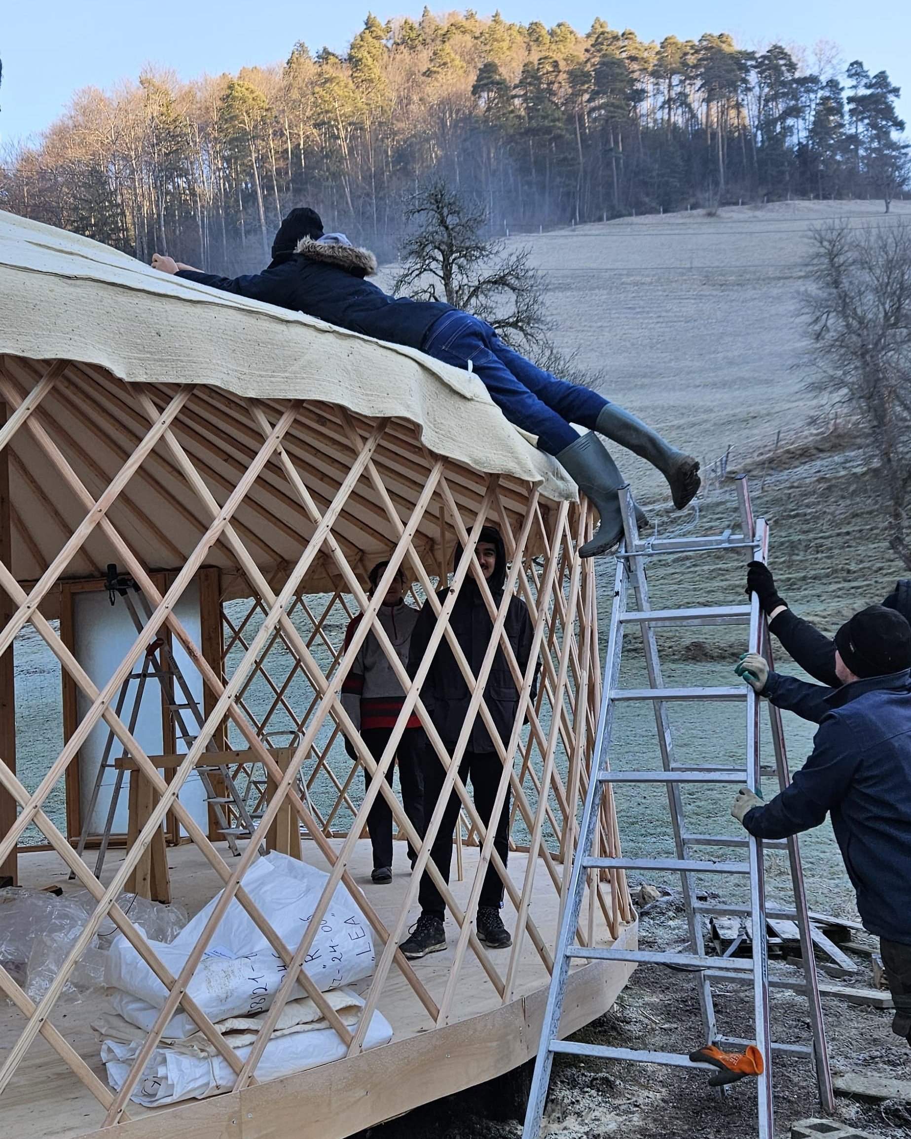 Personen bauen eine Jurte in einer ländlichen Umgebung auf. Einige Menschen stehen auf einer Leiter und befestigen die Dachabdeckung. Eine Person liegt auf der Dachkante. Im Hintergrund sind Bäume und Felder zu sehen.