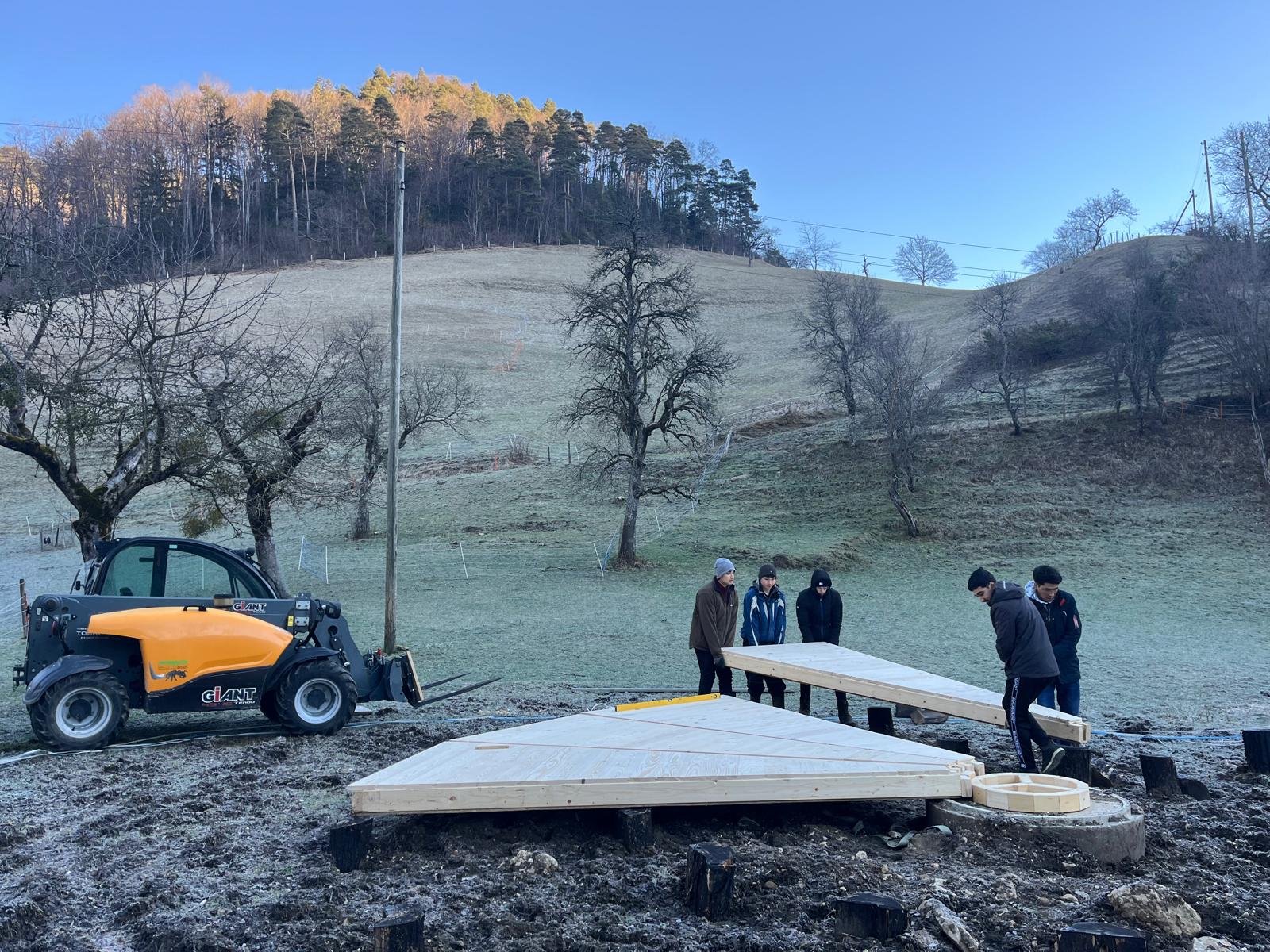 Eine Gruppe von Menschen arbeitet im Freien an einem Bauprojekt. Sie heben Holzelemente auf einem unebenen, grasbewachsenen Gelände. Im Hintergrund sind Bäume und ein hellblauer Himmel sichtbar. Ein kleiner, orangefarbener Bagger befindet sich links