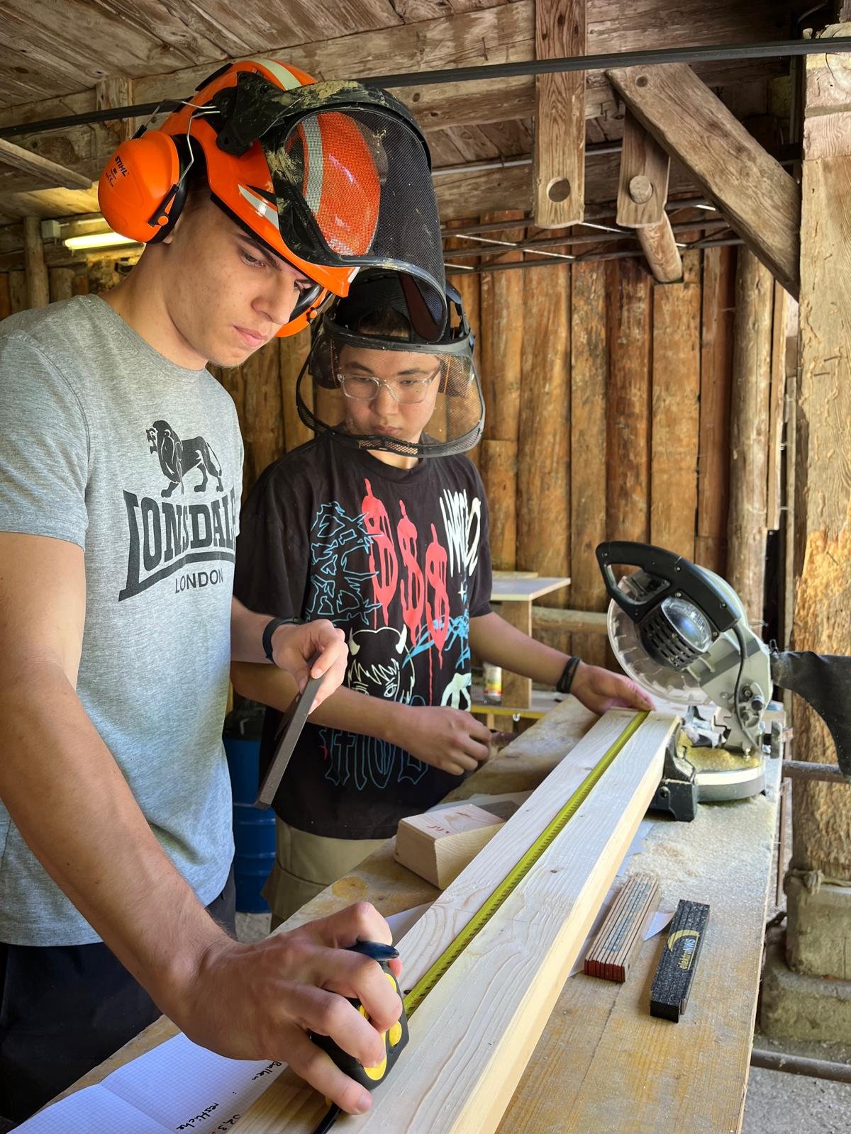 Zwei Männer arbeiten mit Holz in einer Werkstatt. Sie tragen Schutzhelme und Gesichtsschutz. Auf dem Tisch liegt ein Holzbrett mit einem Maßband, und im Hintergrund steht eine Kreissäge.