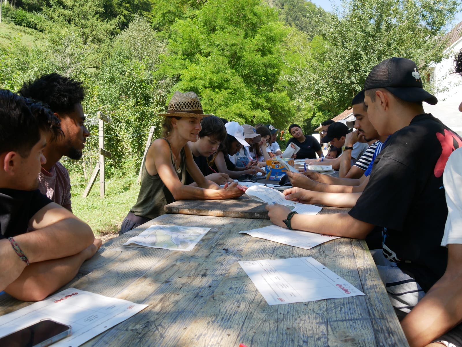 Gruppe junger Menschen sitzt an einem langen Holztisch im Freien, umgeben von Natur. Sie schauen auf Dokumente und diskutieren. Im Vordergrund sind Kartendokumente zu sehen. Die Szenerie wirkt sommerlich und entspannt.