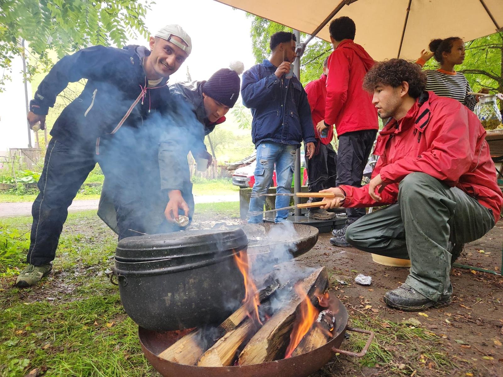 Gruppe von Personen bei einem Picknick im Freien, mehrere in Jacken, eine bückt sich über einen Lagerfeuertopf, während eine andere mit einem Stöckchen im Feuer rührt. Rauch steigt auf, und einige lachen. Im Hintergrund stehen weitere Personen unter