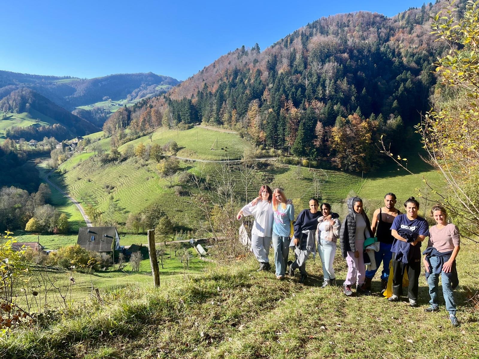 Gruppe von Menschen steht auf einem Hügel mit Aussicht auf eine malerische Landschaft. Im Hintergrund sind grüne Hügel und Wälder mit herbstlichen Farben zu sehen. Der Himmel ist klar und blau.