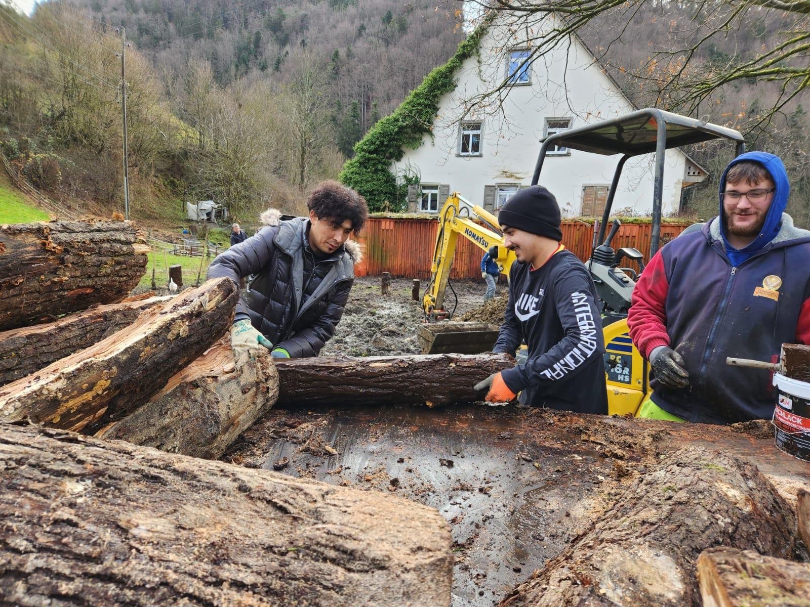 Drei Personen arbeiten im Freien mit Baumstämmen, während im Hintergrund ein Bagger zu sehen ist. Sie tragen Winterkleidung und Handschuhe. Die Umgebung ist ländlich mit Bäumen und einem Haus im Hintergrund.