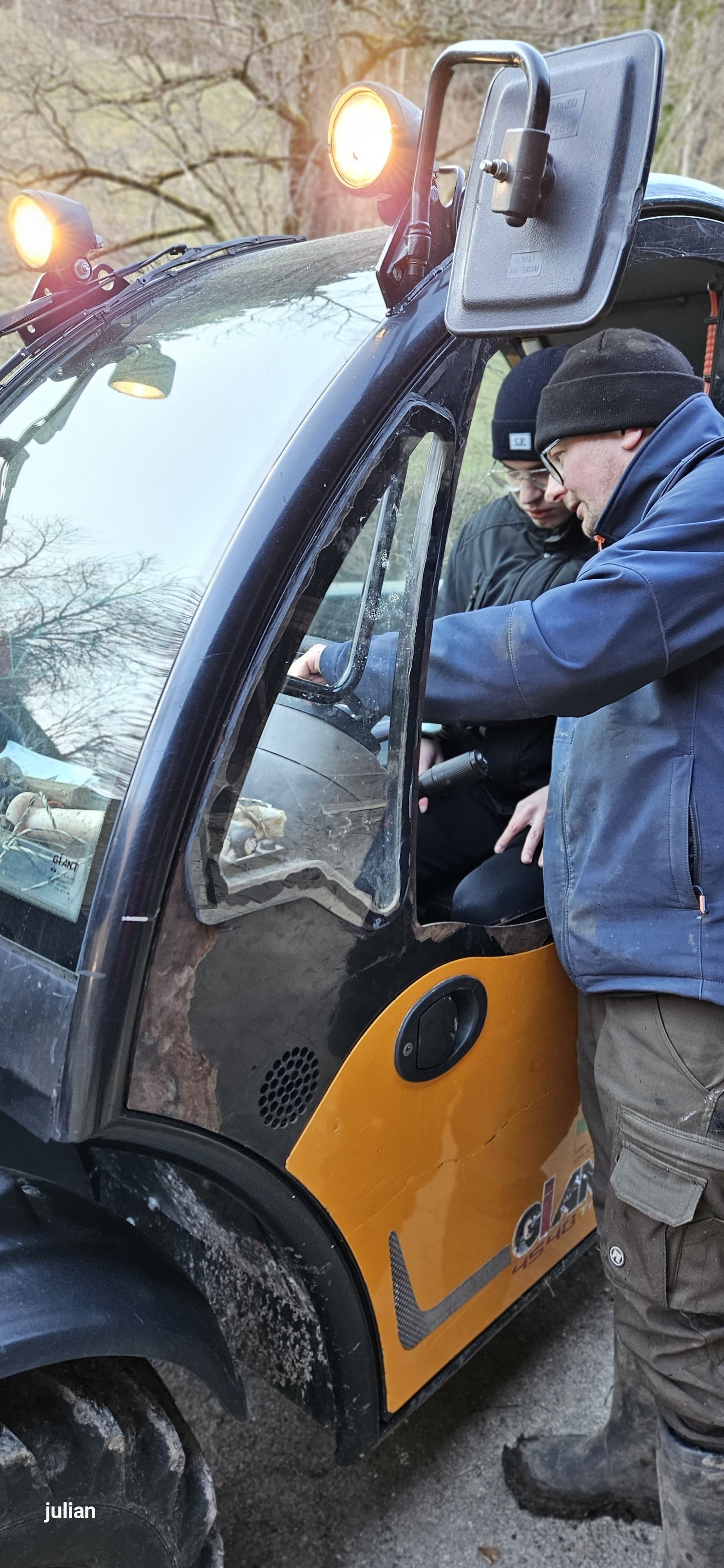 Zwei Personen stehen neben einem kleinen gelben Fahrzeug mit offener Tür. Eine Person zeigt auf das Armaturenbrett. Im Hintergrund sind Bäume sichtbar. Das Fahrzeug hat leuchtende Scheinwerfer oben.
