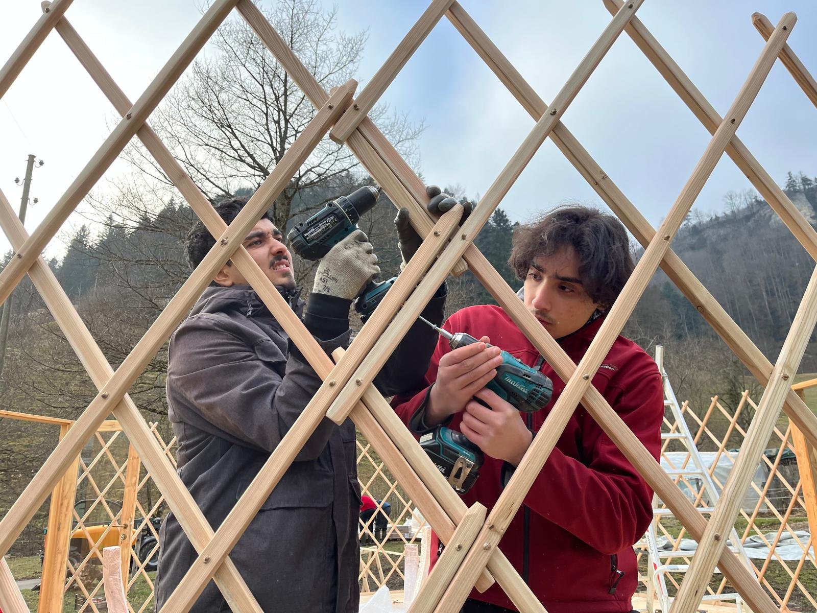 Zwei Menschen arbeiten mit Akkuschraubern an einer hölzernen Struktur im Freien, umgeben von einer malerischen Waldlandschaft.
