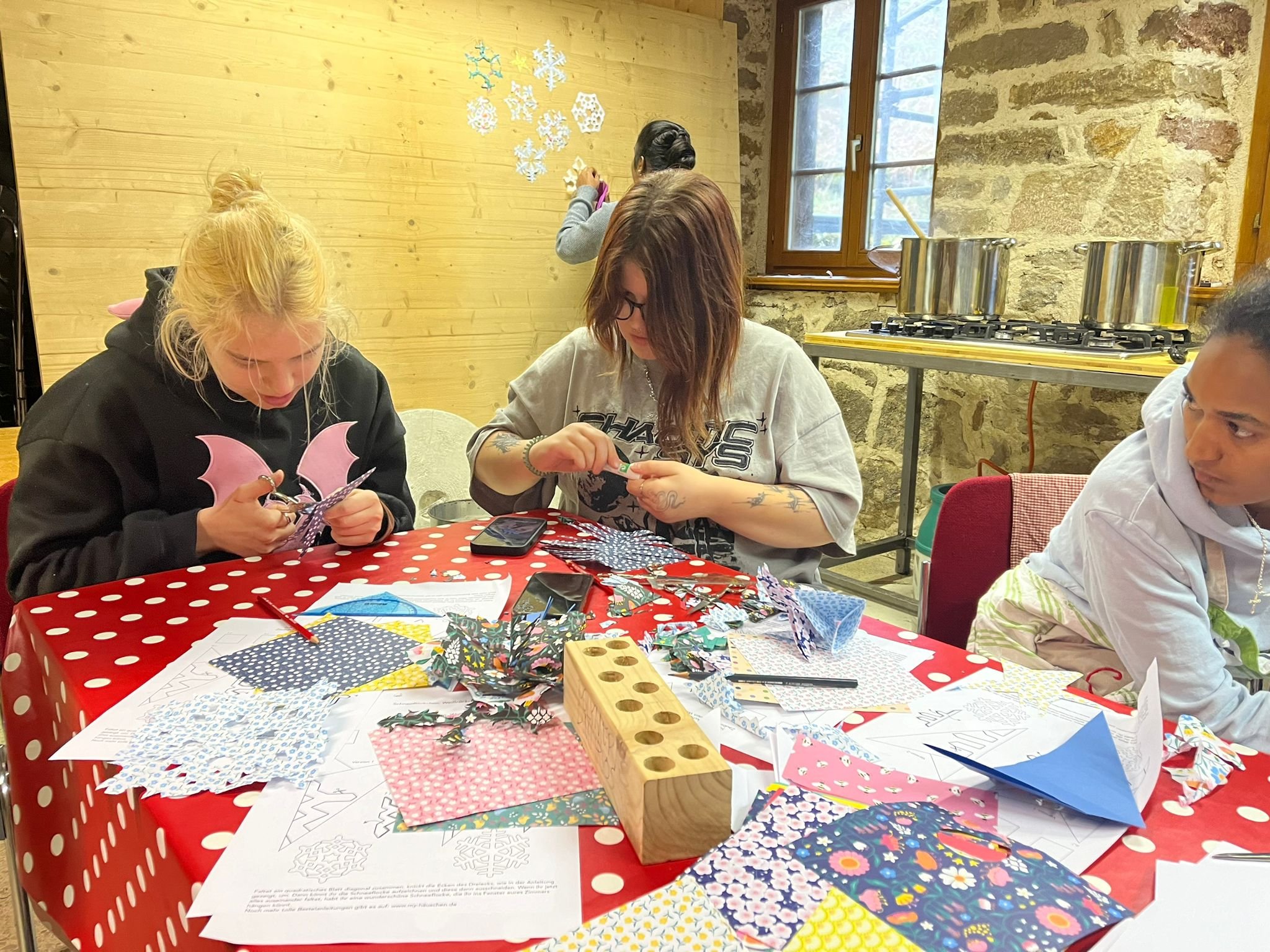 Drei Personen basteln Schnee- und Blumenmuster aus Papier an einem Tisch mit rot-weiß gepunkteter Tischdecke in einem rustikalen Raum.